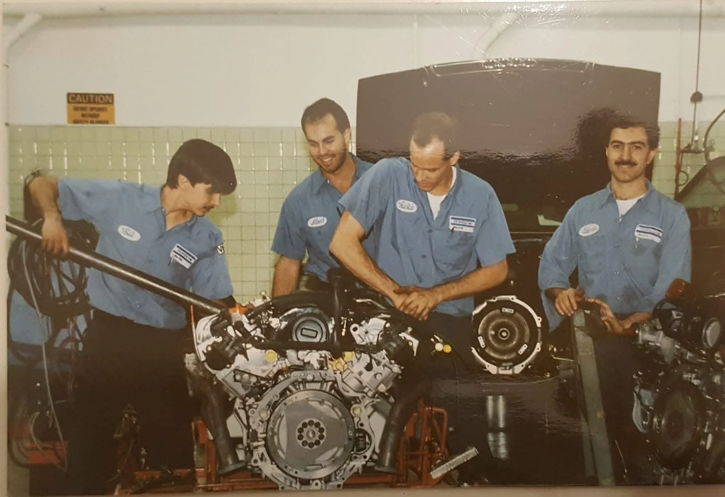 Four mechanics in blue uniforms working on car engines in a garage.