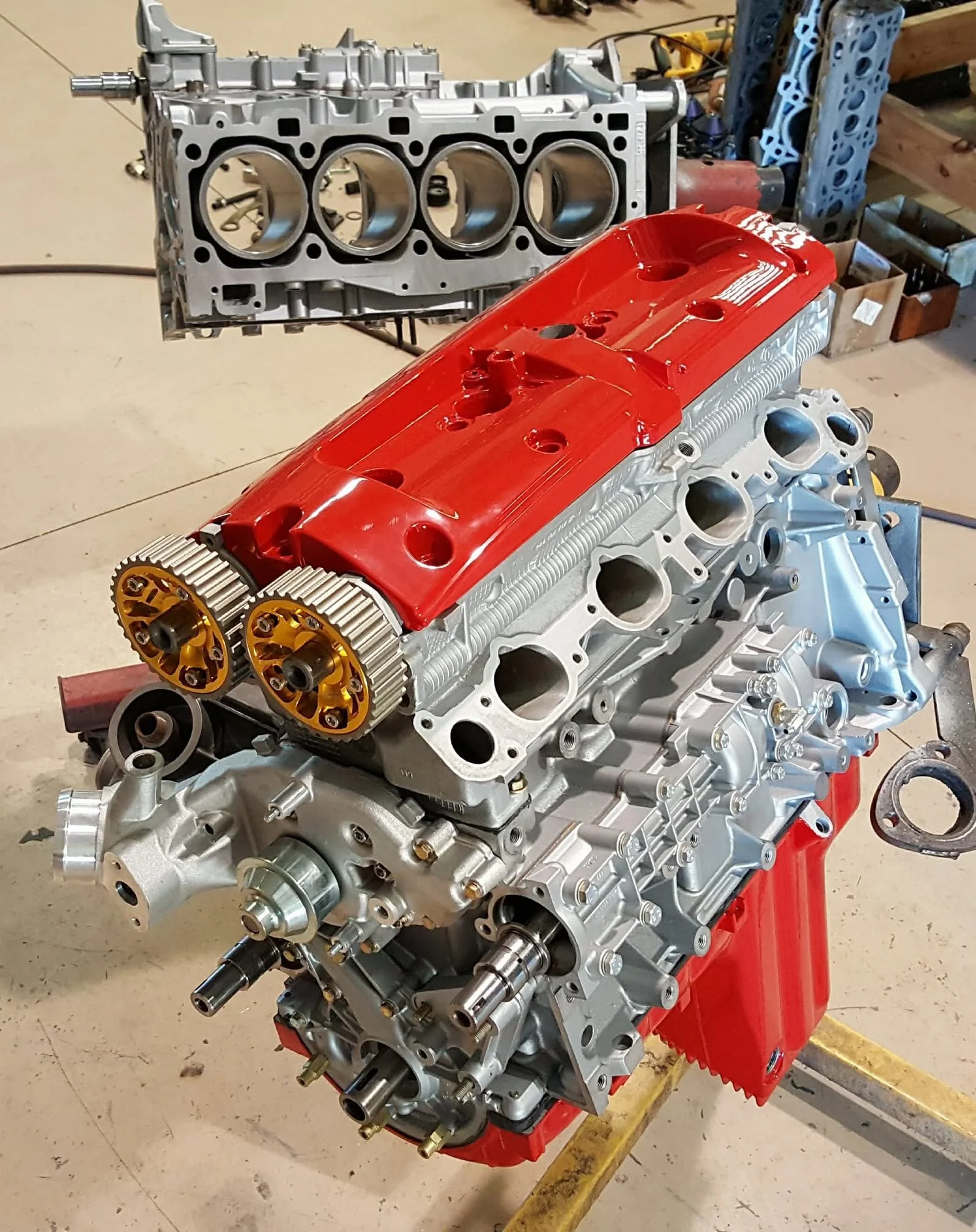 Disassembled car engine with red valve cover and visible internal components on a workshop floor.