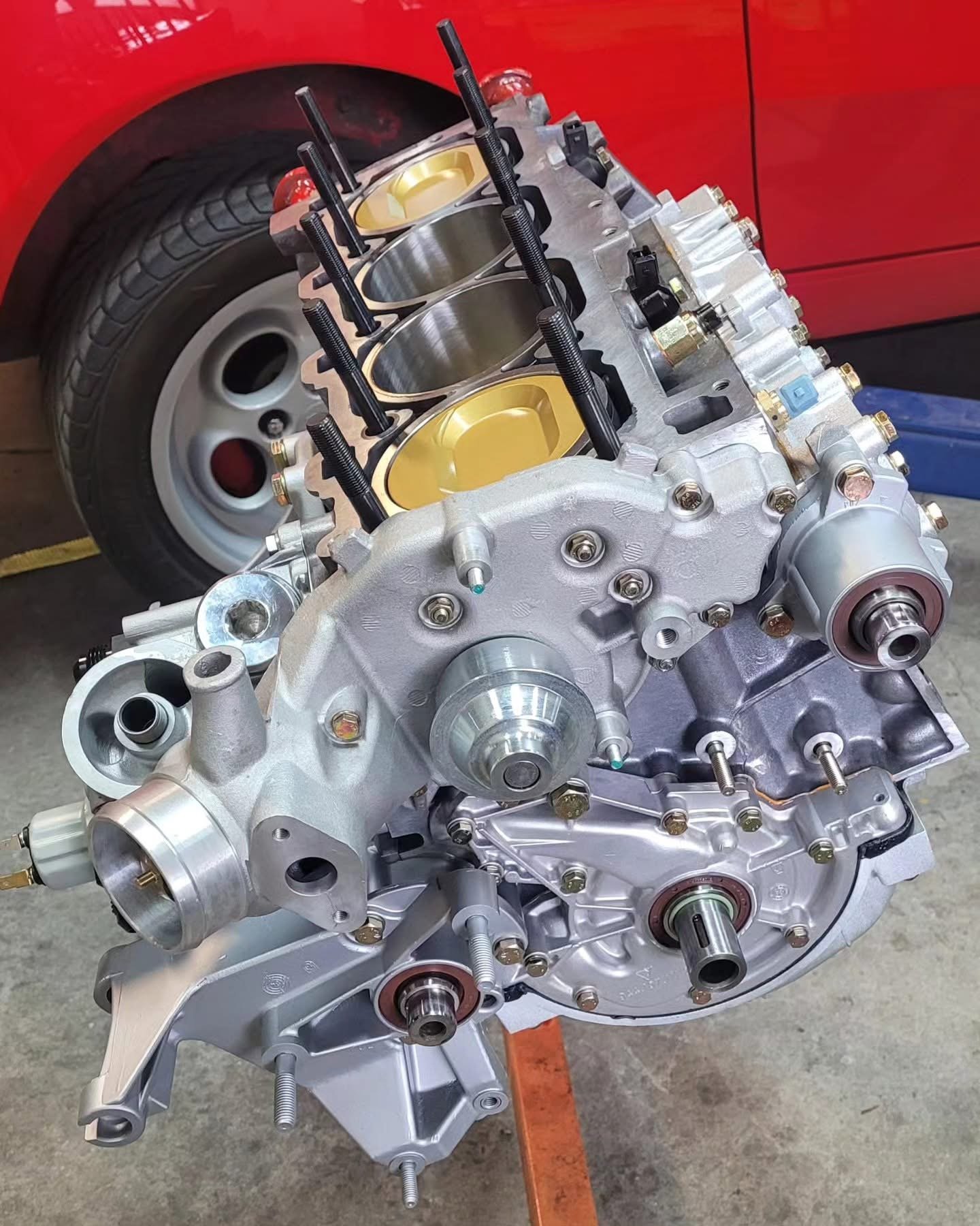 Car engine block with exposed pistons and surrounding components, positioned next to a red vehicle with a visible tire.