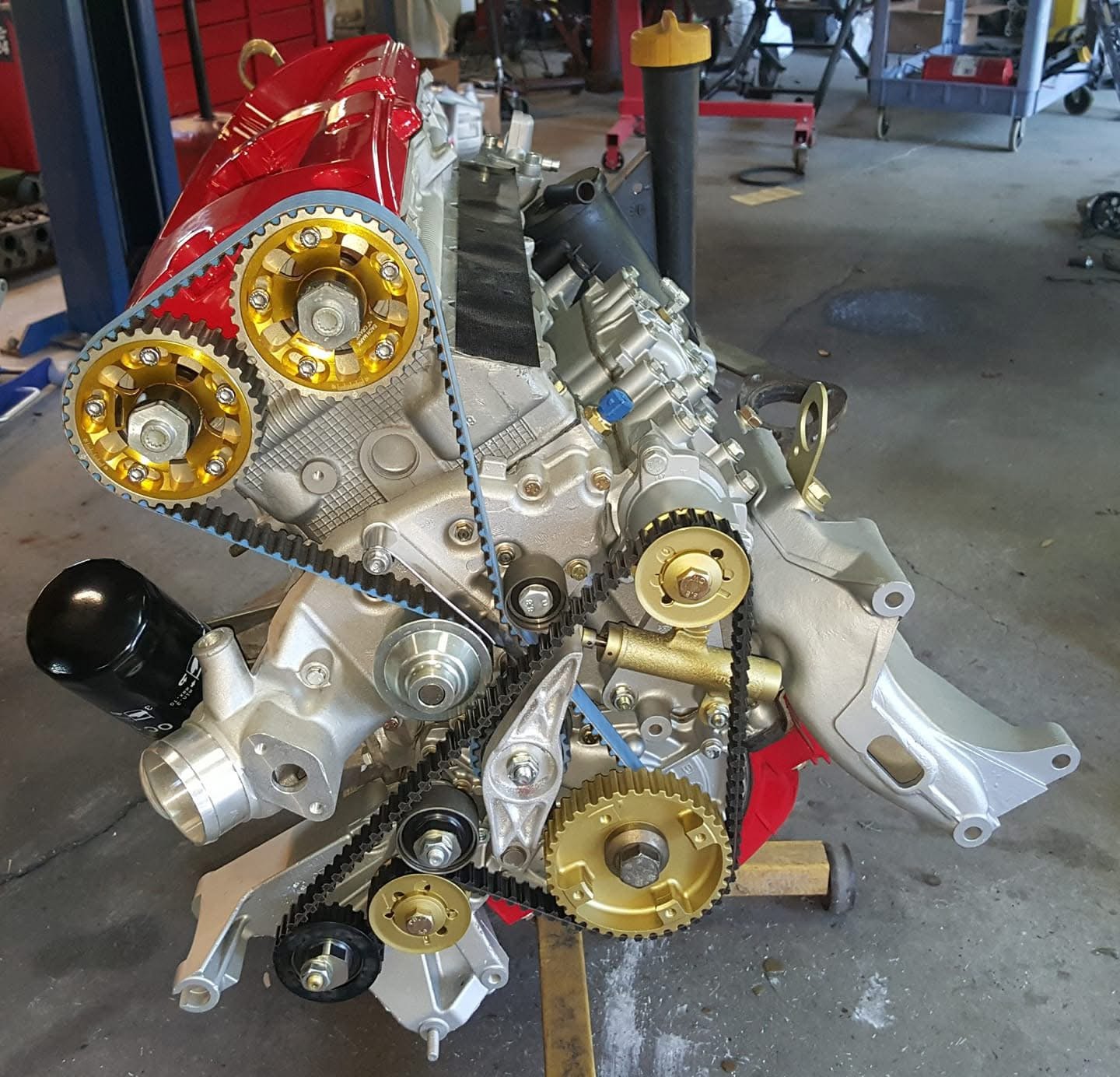 High-performance car engine with red valve cover and gold timing gears, mounted on a stand in a garage.