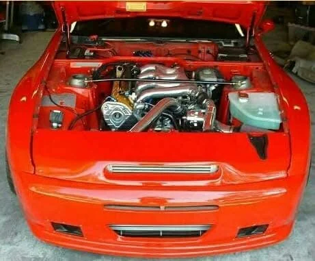 Front view of a red car with hood open, showcasing a rotary engine. The engine bay is clean and well-organized, highlighting the vehicle's mechanical components.