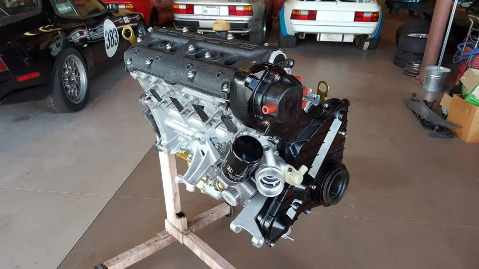 A car engine mounted on a stand inside a garage, surrounded by several parked cars and tools.