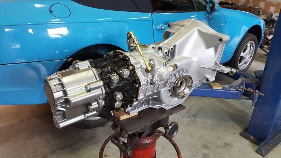 A freshly refurbished car transmission on a stand in an auto shop, with a blue sports car in the background.