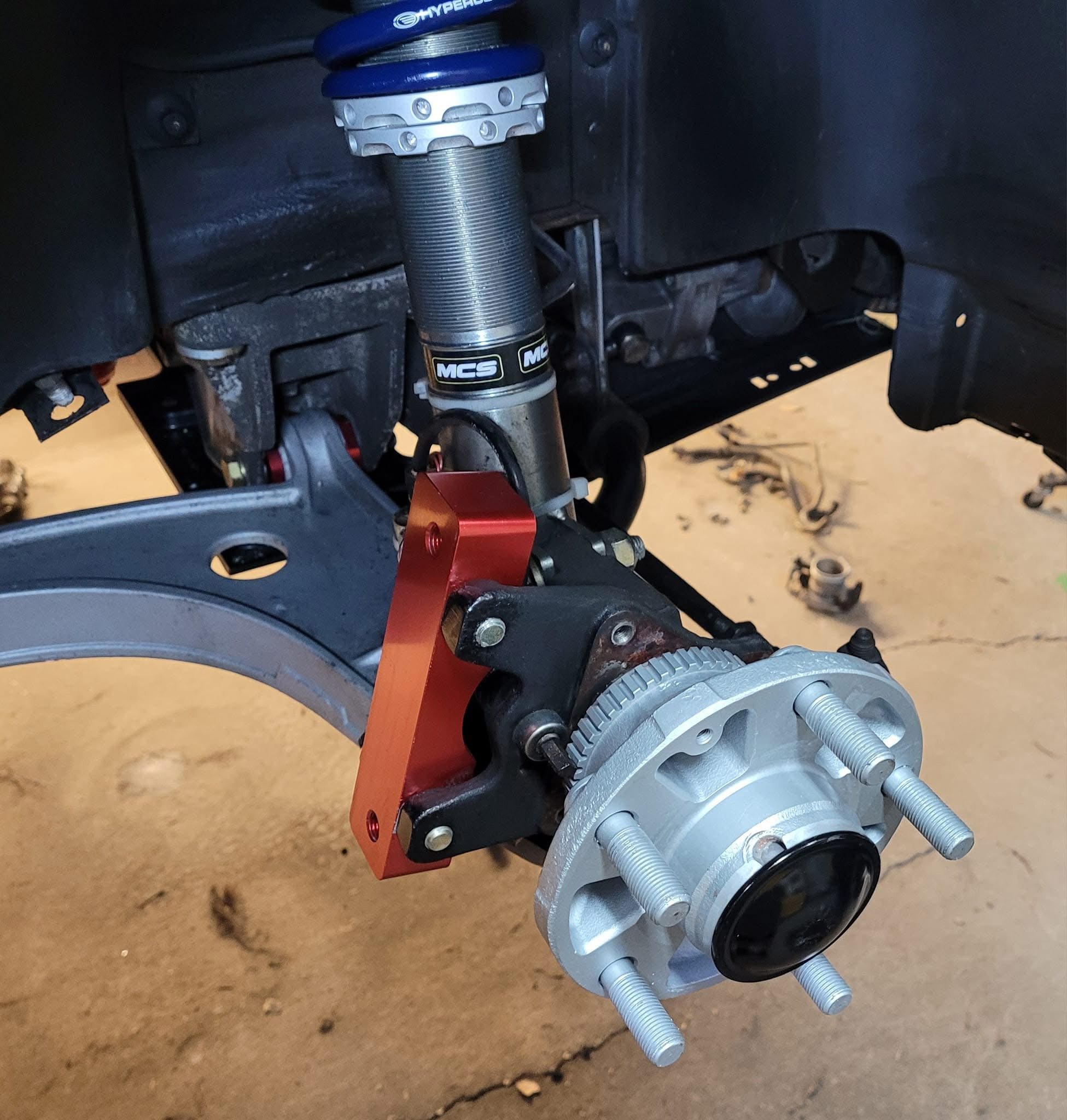 Close-up of a car wheel assembly showcasing a performance suspension setup, including a coilover with a blue top and a silver cylindrical component, alongside a red bracket and silver hub with five lug bolts.