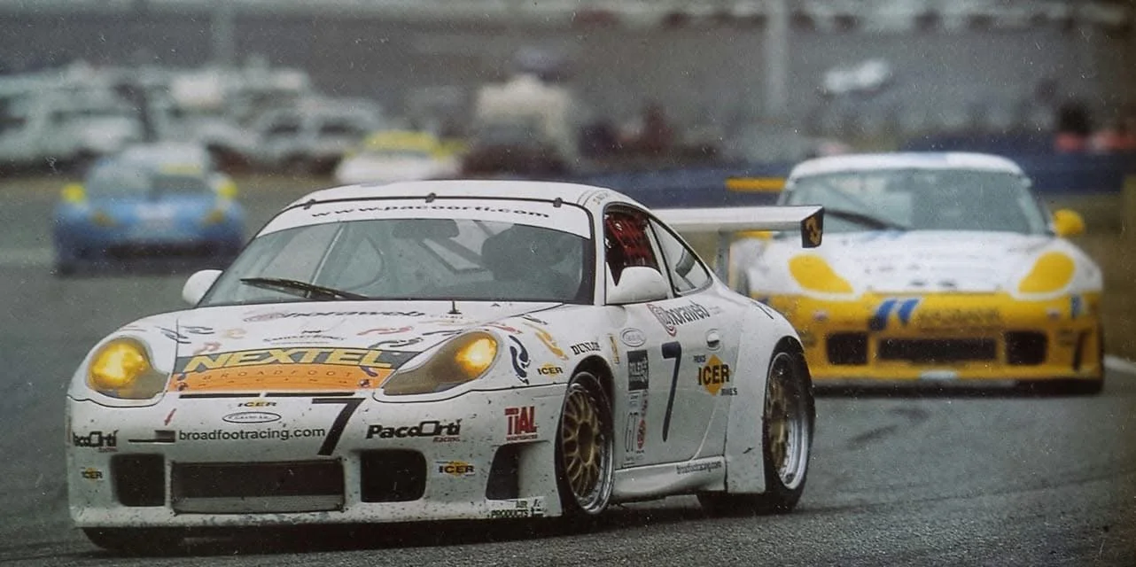 Racing cars on a wet track during a motorsport event, with focus on a white Porsche in the foreground and a yellow and blue car behind.