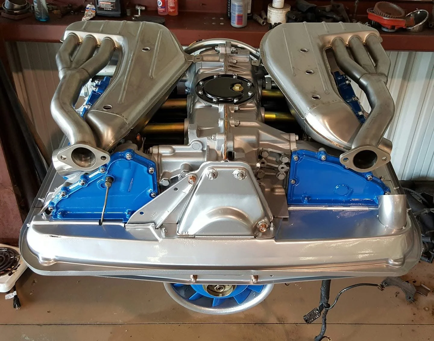 Porsche flat-six engine with dual exhaust manifolds and blue engine components in a garage.