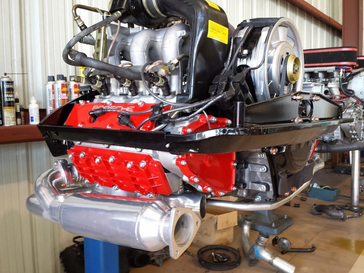 Porsche air-cooled engine, partially disassembled, in a workshop with tools and containers in the background.