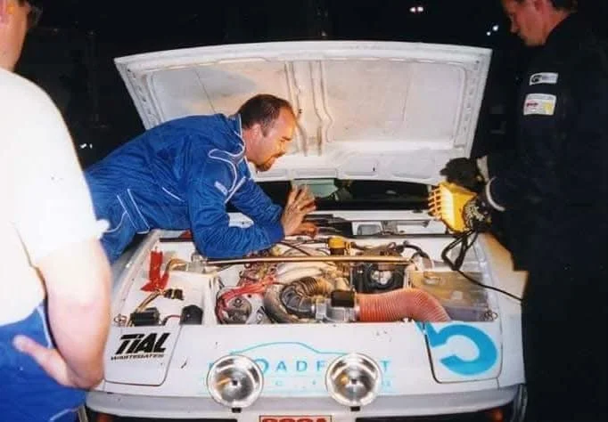 Mechanics inspecting the engine of a rally car during a night event. One person is examining while another holds a work light, illuminating the engine bay. The car has multiple sponsor stickers and rally lights.