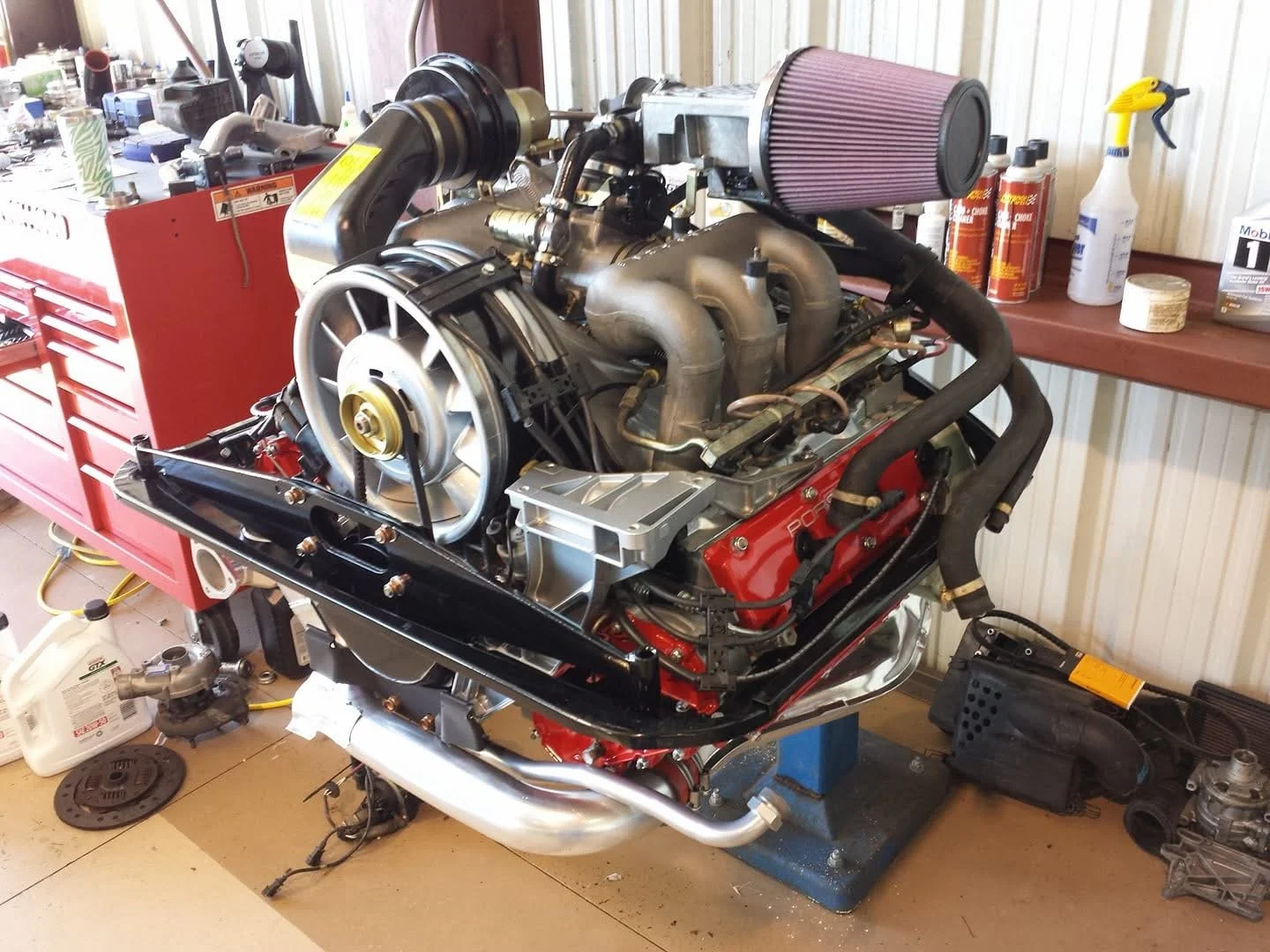 Porsche 911 engine on a stand in a workshop with tools and supplies in the background.