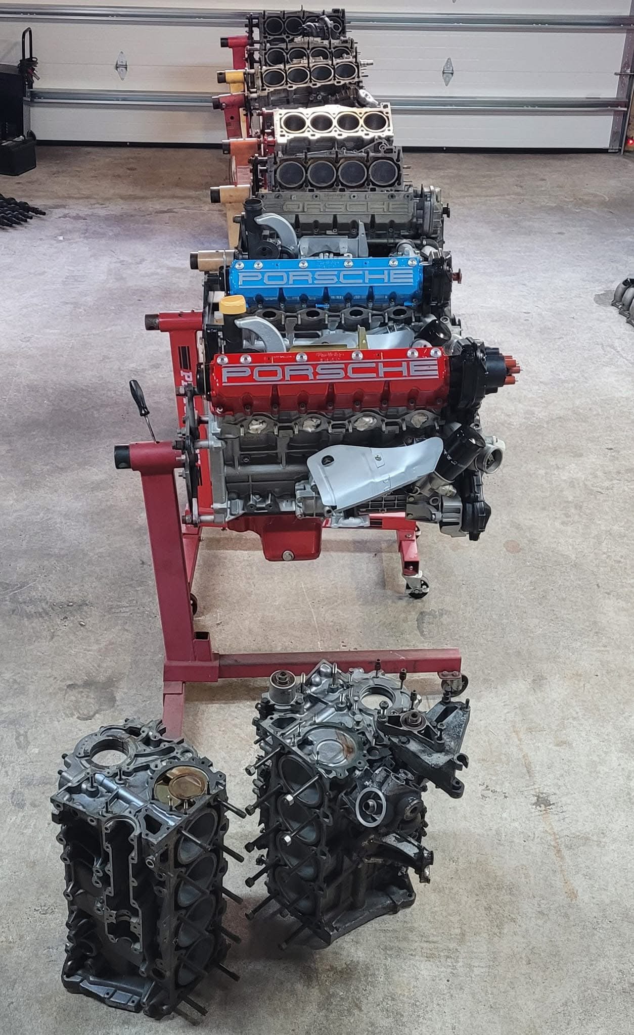 Several Porsche engine blocks lined up in a garage on stands, with some disassembled parts on the floor.