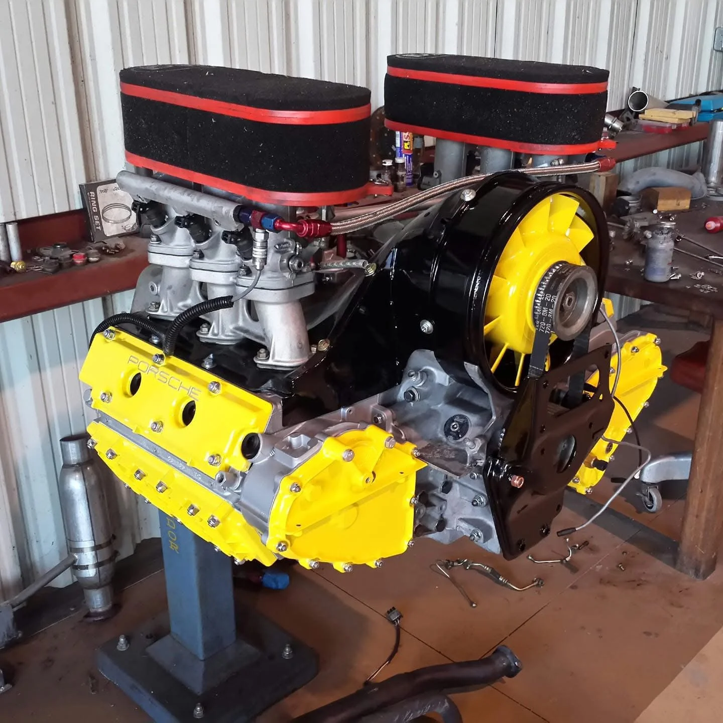 Restored Porsche engine with yellow valve covers and air filters on a stand in a garage.