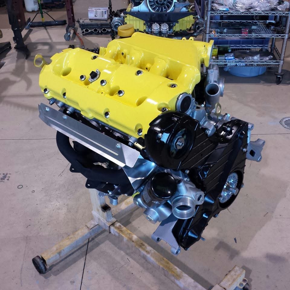 Bright yellow Porsche 944 engine on a stand in a workshop setting, with various mechanical parts visible in the background.