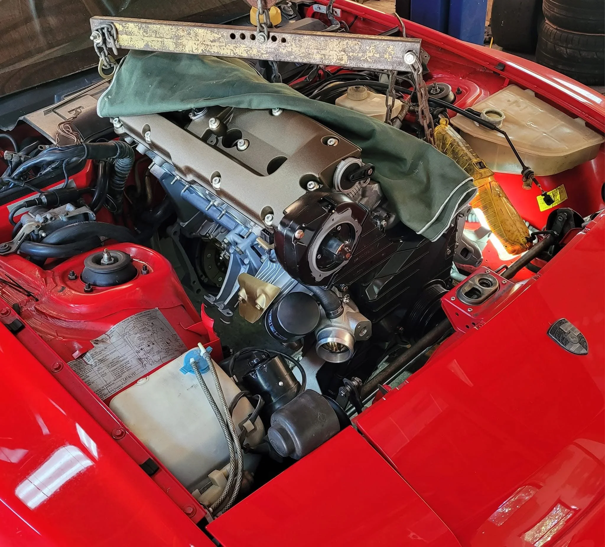 Car engine installation in a red vehicle's engine bay, with a hoist holding the engine. A green cloth partially covers the engine.