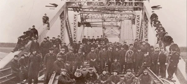 Black and white photograph of many soldiers gathered on a ship's deck, some sitting on the rails, others standing, with the ship's structure and rigging visible overhead.