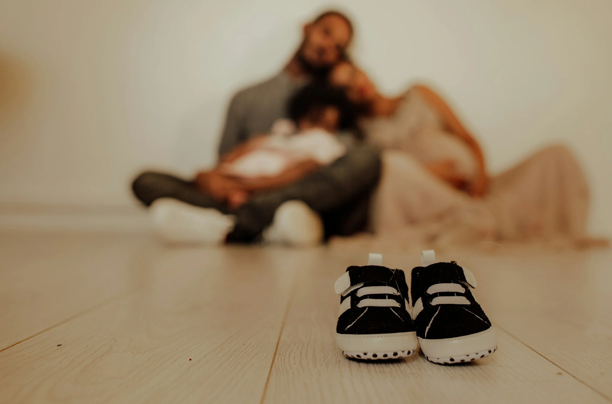 Pair of tiny black and white baby shoes on a light wooden floor, out of focus family of three with the parents holding a newborn in the background.