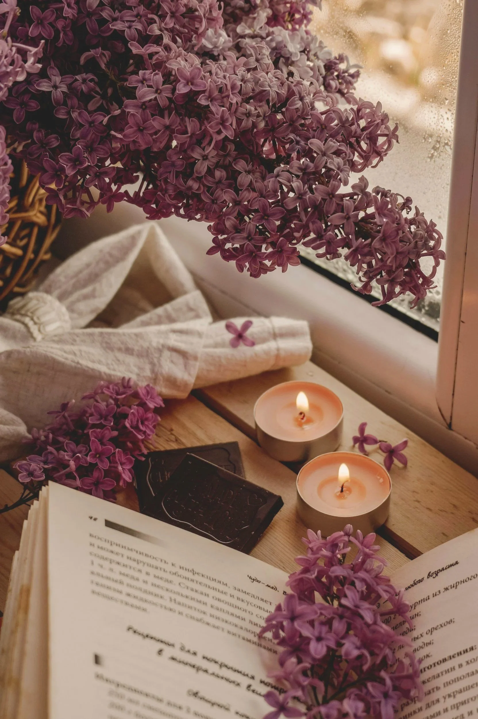 Purple lilac flowers on a windowsill with two lit candles, a wicker basket, an open book with text, some cloth, and a black box.