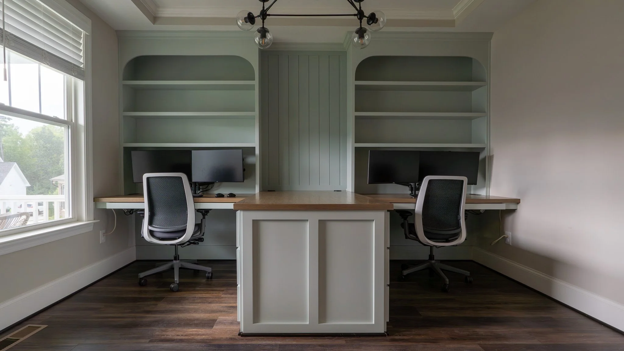 Home office with two computer stations, swivel chairs, built-in shelves, and large window.
