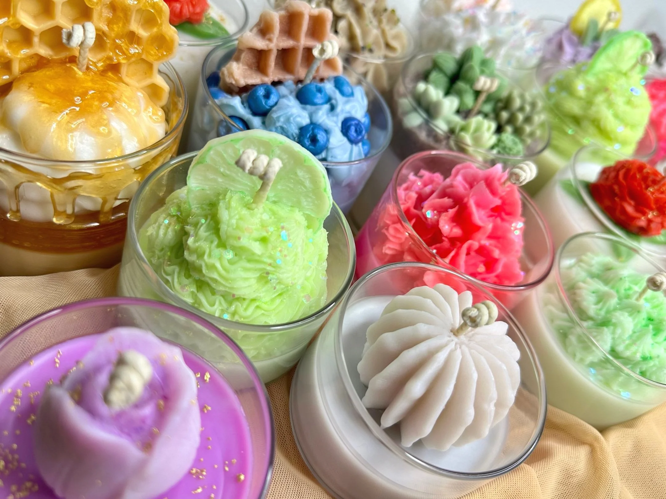 Assorted decorative candles in glass containers, shaped like desserts and fruits, featuring vibrant colors such as green, pink, purple, and white, with textured wax designs resembling whipped cream, waffles, and honeycombs.