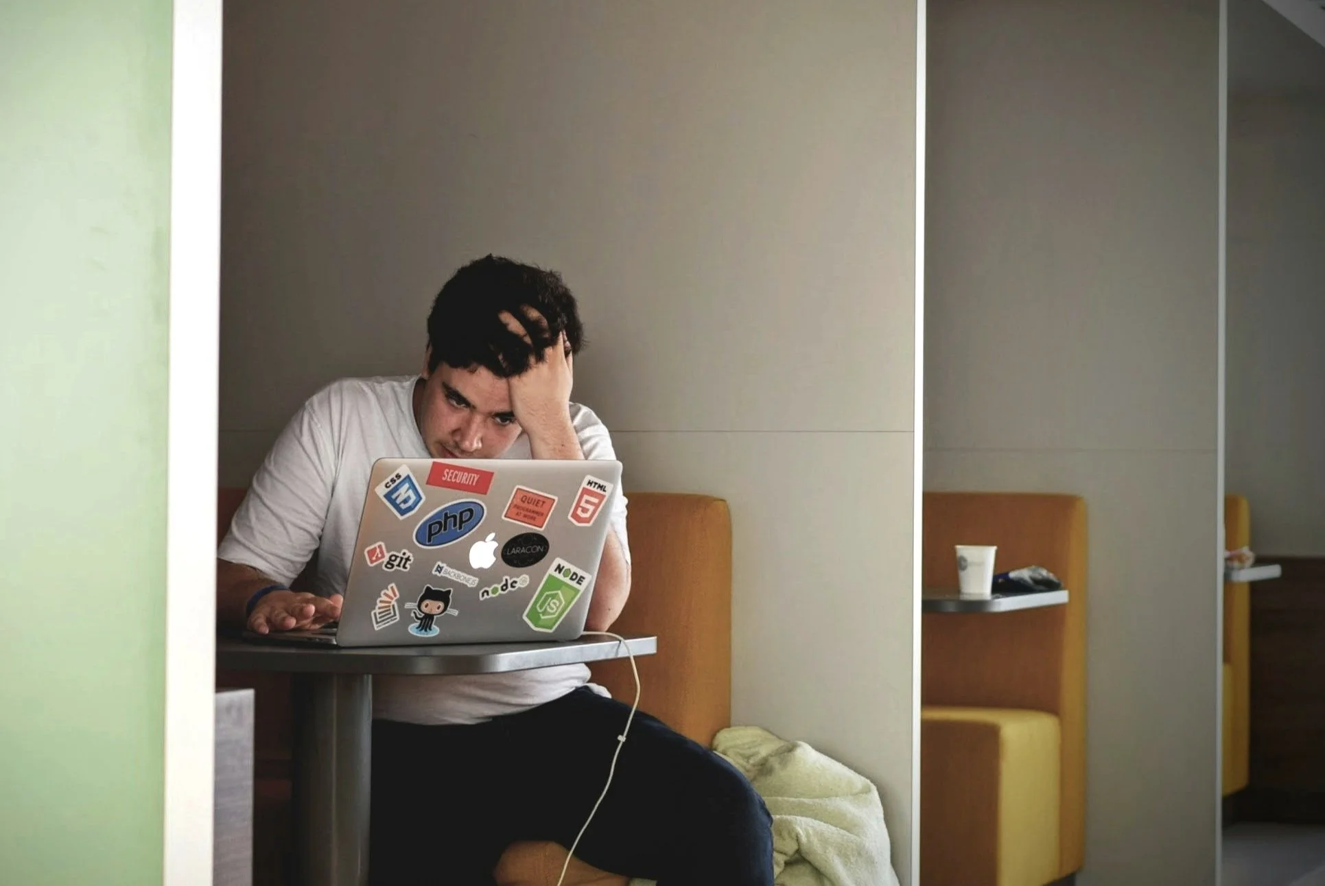 A person in a white t-shirt sitting at a table with an open laptop covered in tech and programming stickers, looking stressed with hand on forehead.