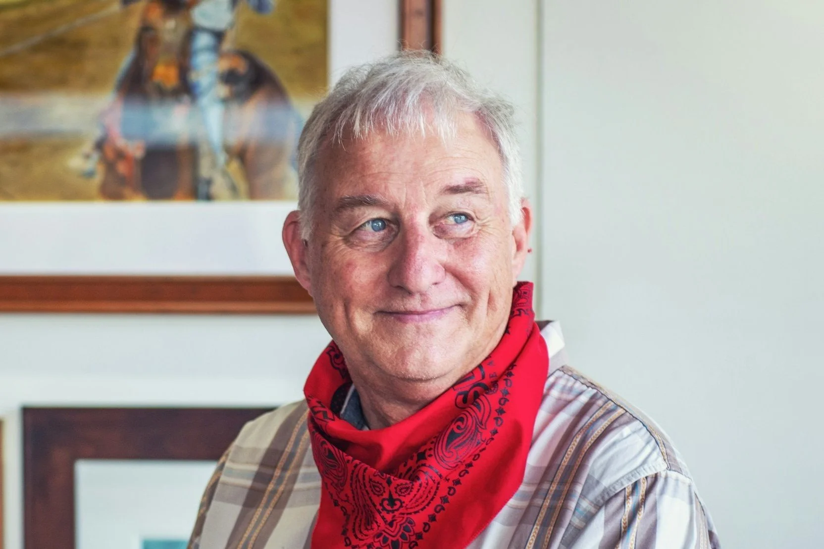 A smiling elderly man with gray hair and blue eyes, wearing a red bandana around his neck and a plaid shirt, indoors with a framed picture of a cowboy riding a horse in the background.