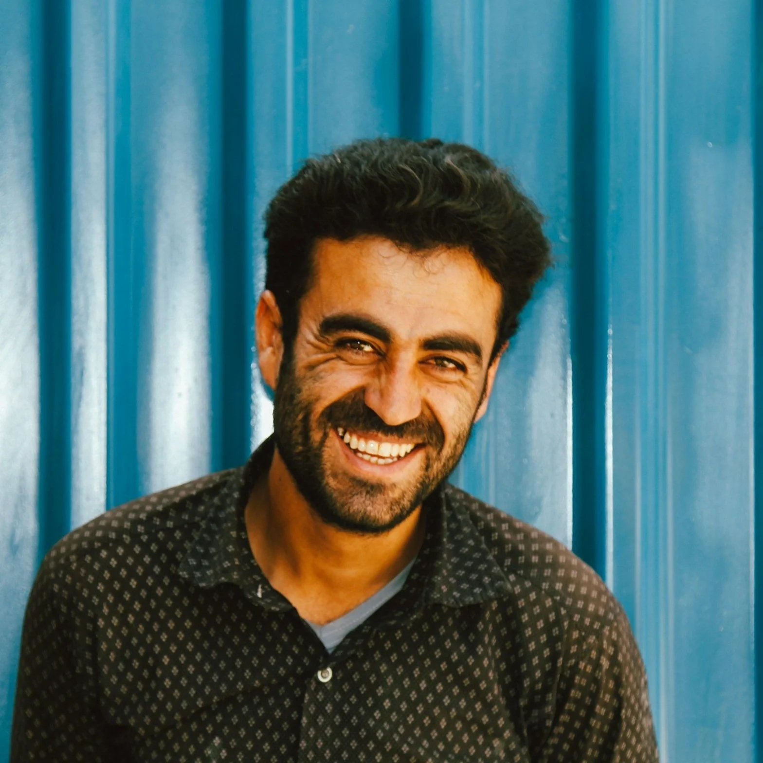 A man with dark hair and a beard smiling in front of a blue metal wall.