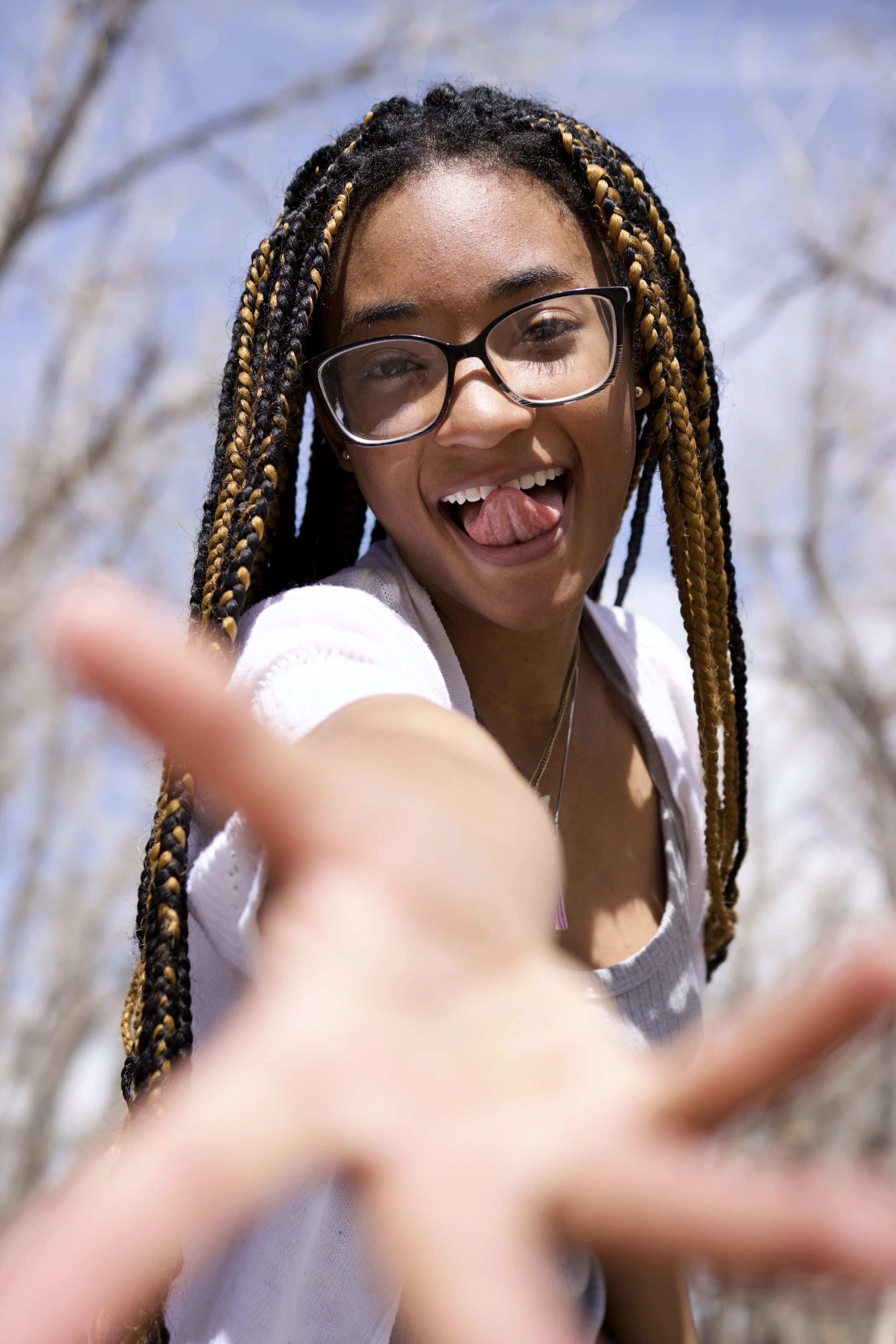 Young woman with braided hair and glasses making a playful gesture outdoors.