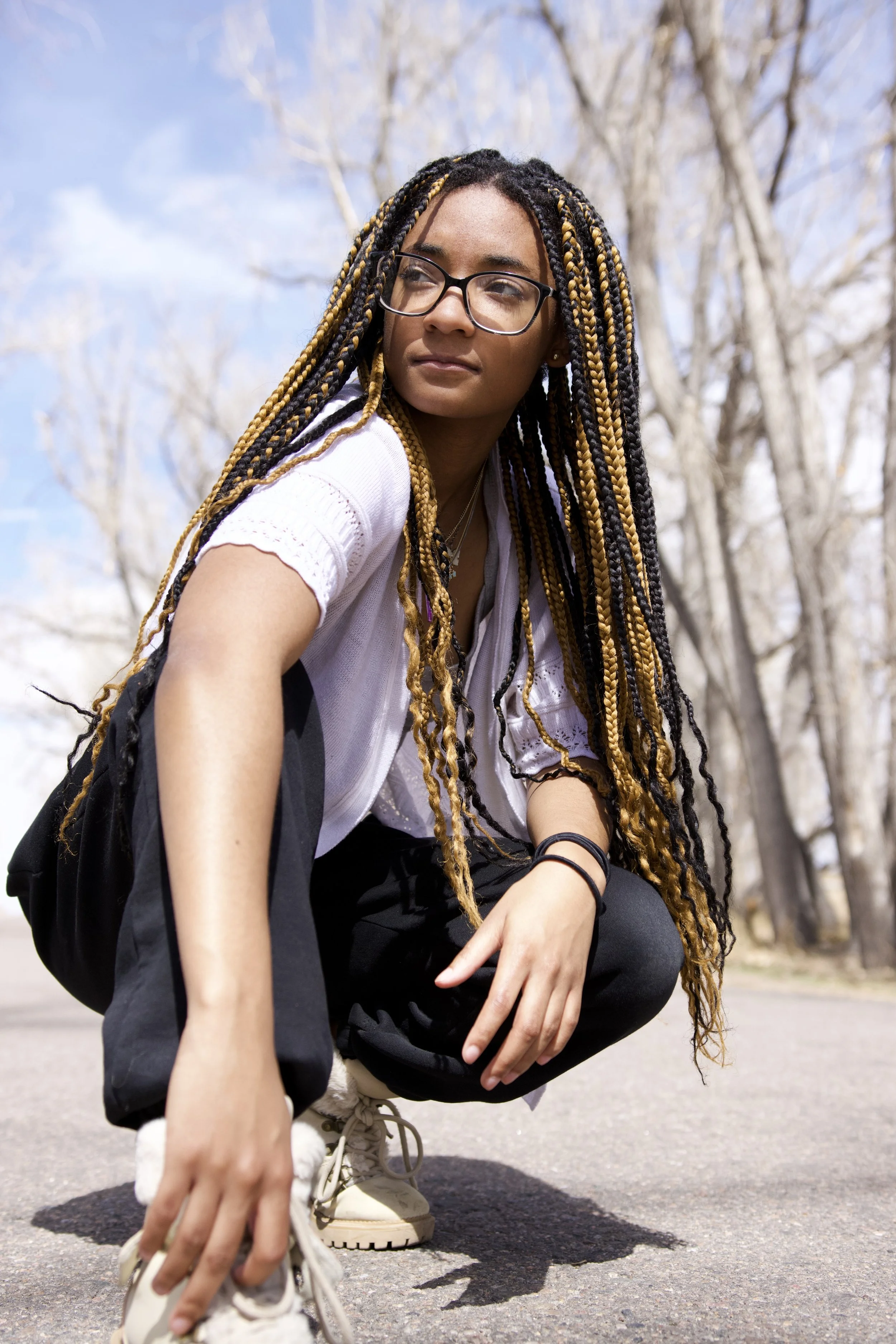 A young woman with glasses and braided hair crouching outdoors on a paved path, with leafless trees in the background and a partly cloudy sky.
