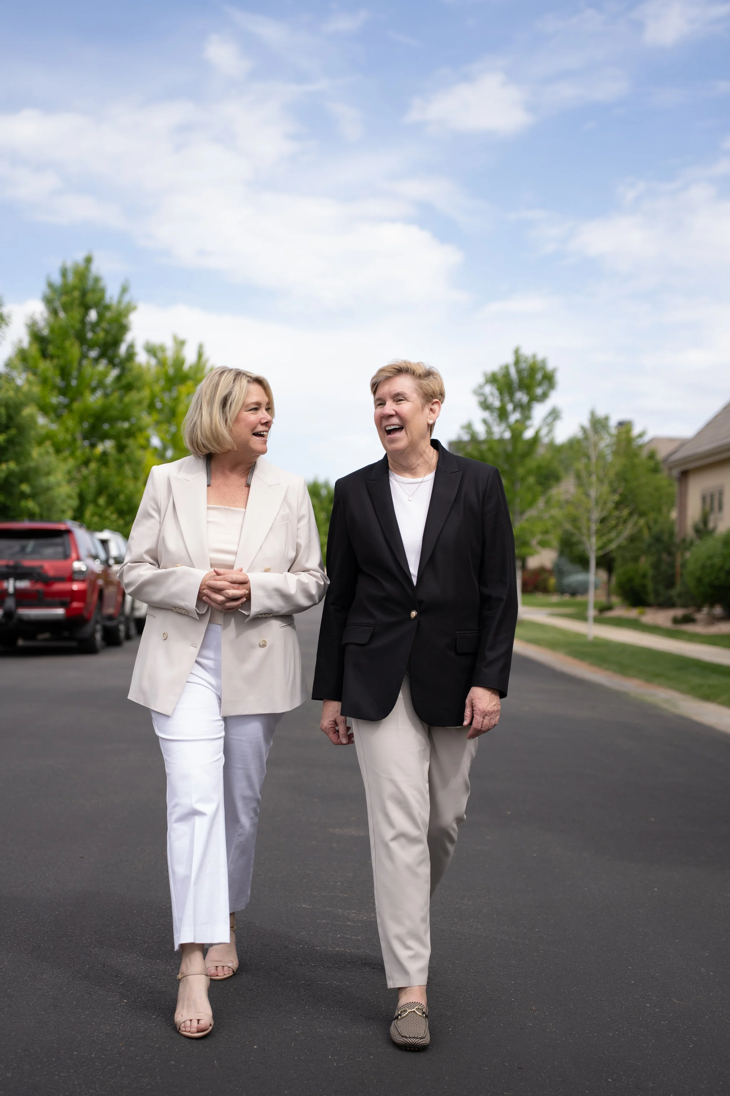 Two women walking and talking on a suburban street, smiling, dressed in business casual attire, with trees and houses in the background.