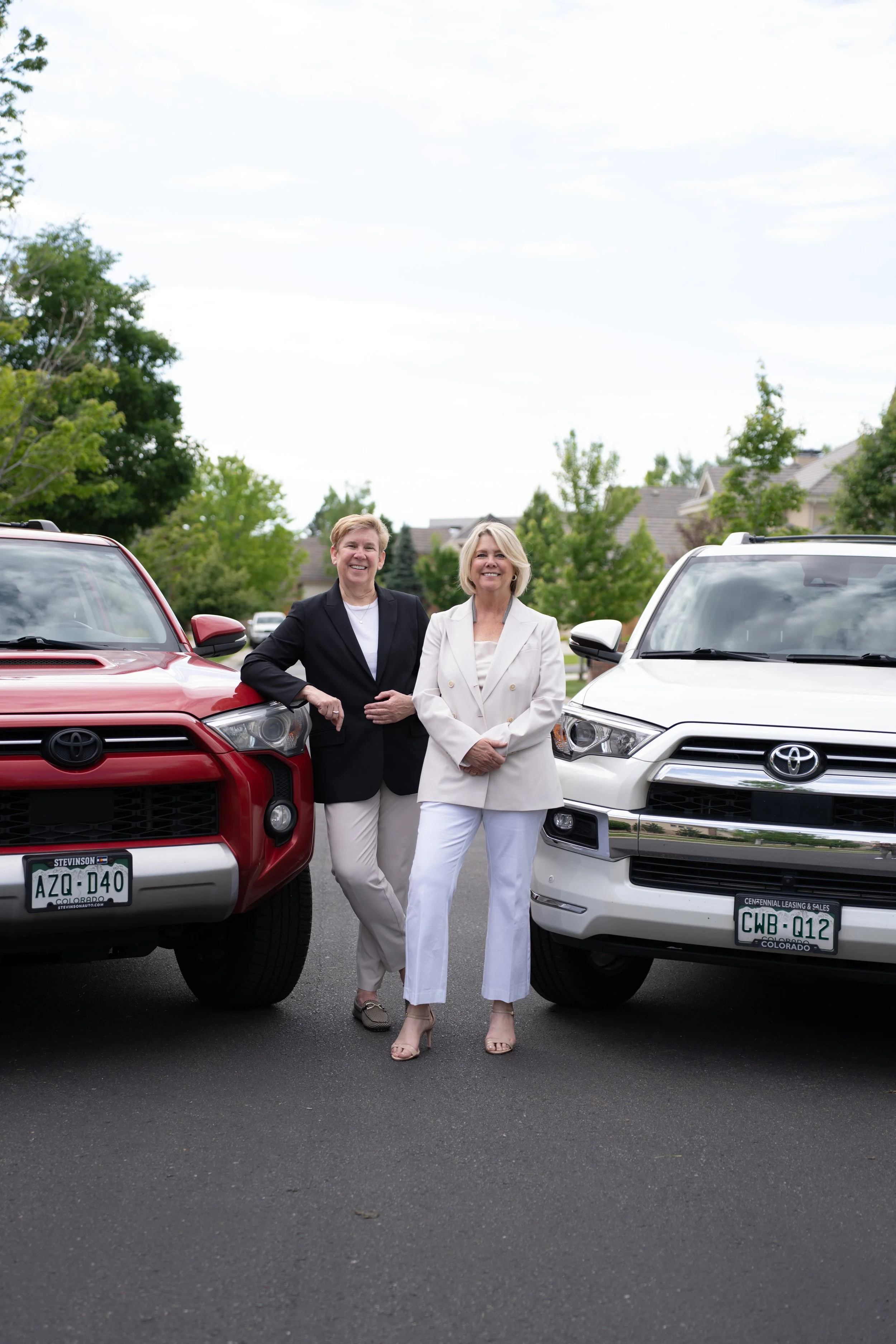 Two women standing between two parked Toyota SUVs in a neighborhood driveway, smiling at the camera.