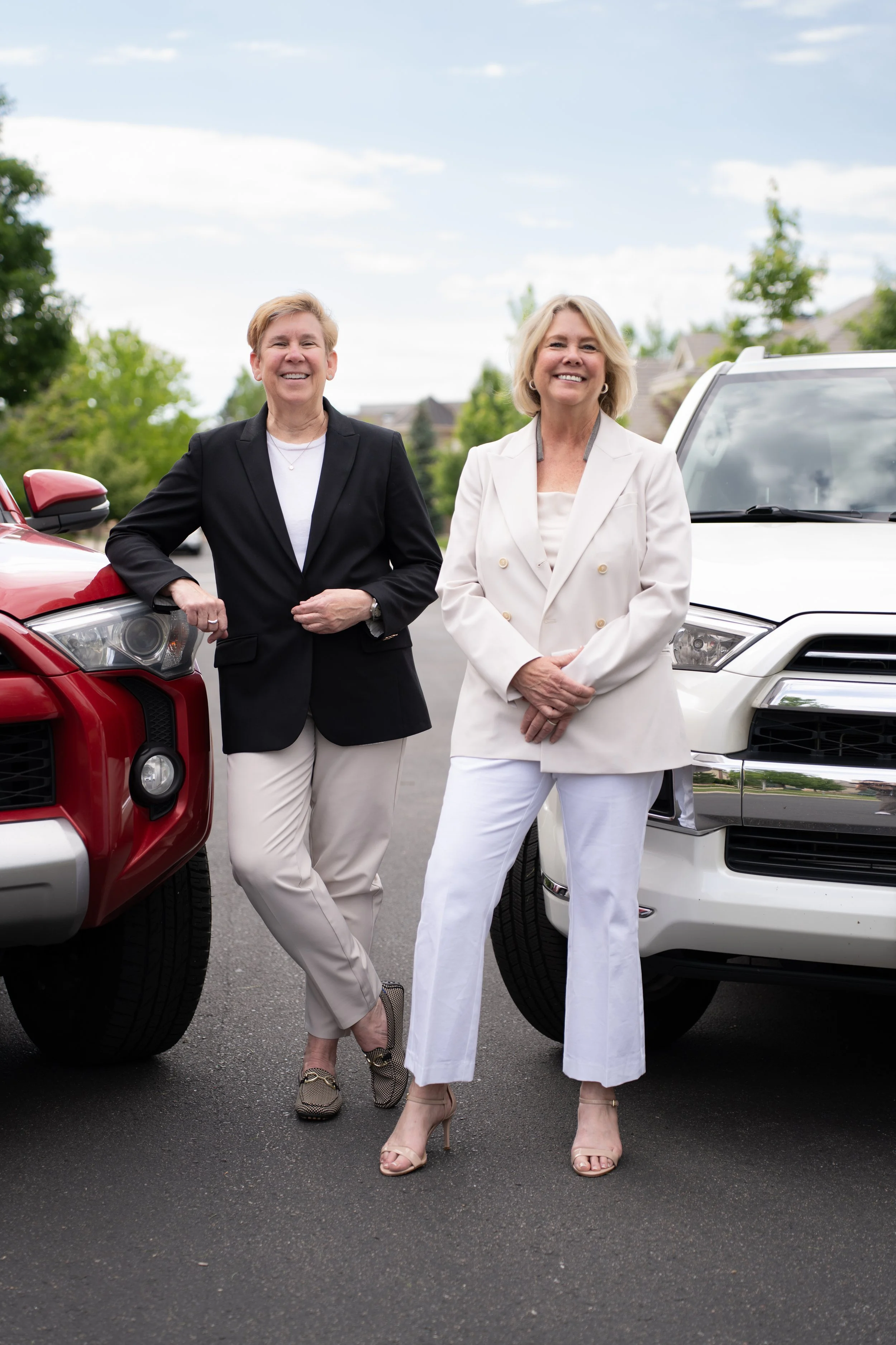 Two women standing outdoors in front of parked cars, smiling, dressed in business casual attire, with trees and houses in the background.