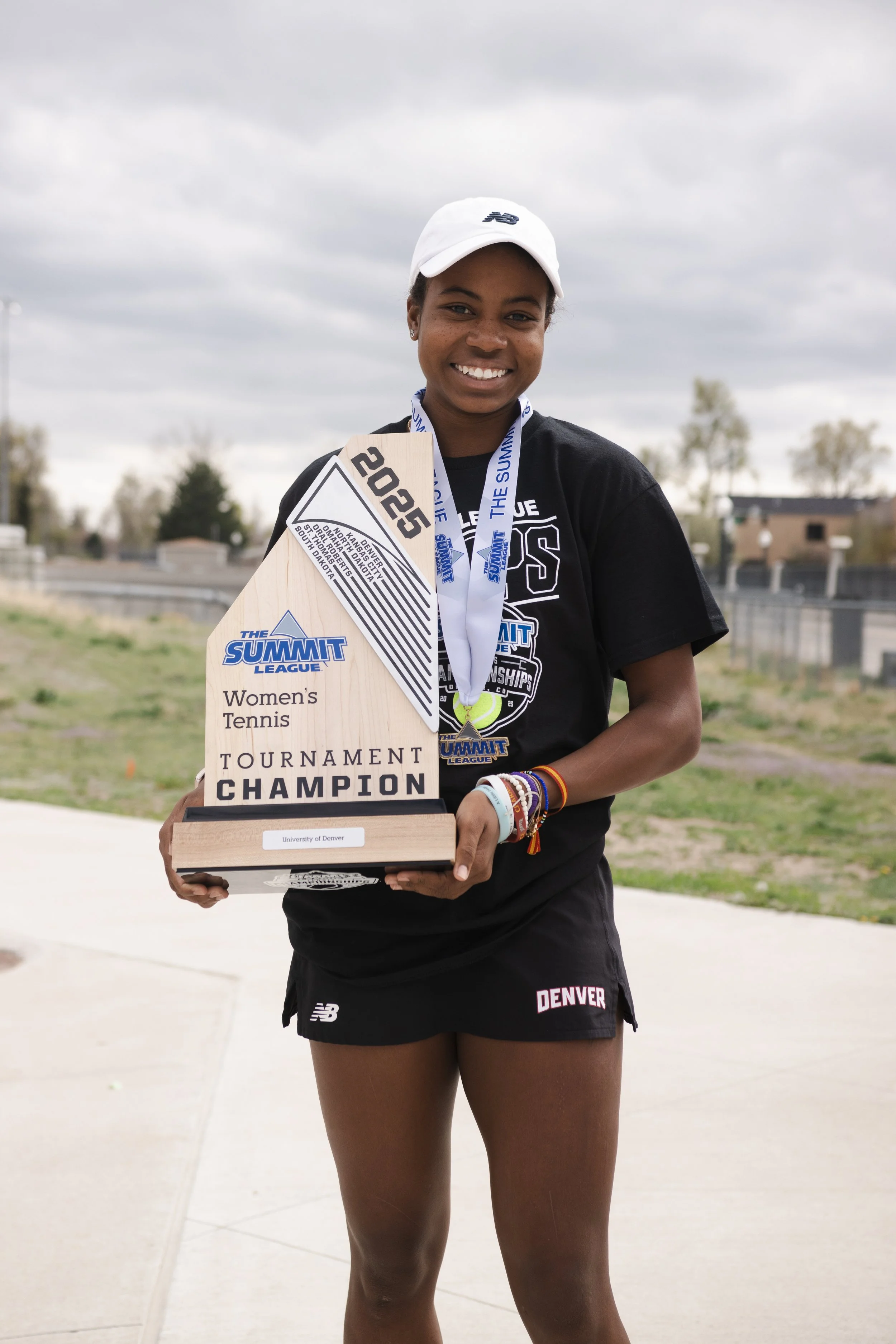 A young woman in athletic attire holding a trophy and wearing medals, smiling at the camera outdoors overcast sky.