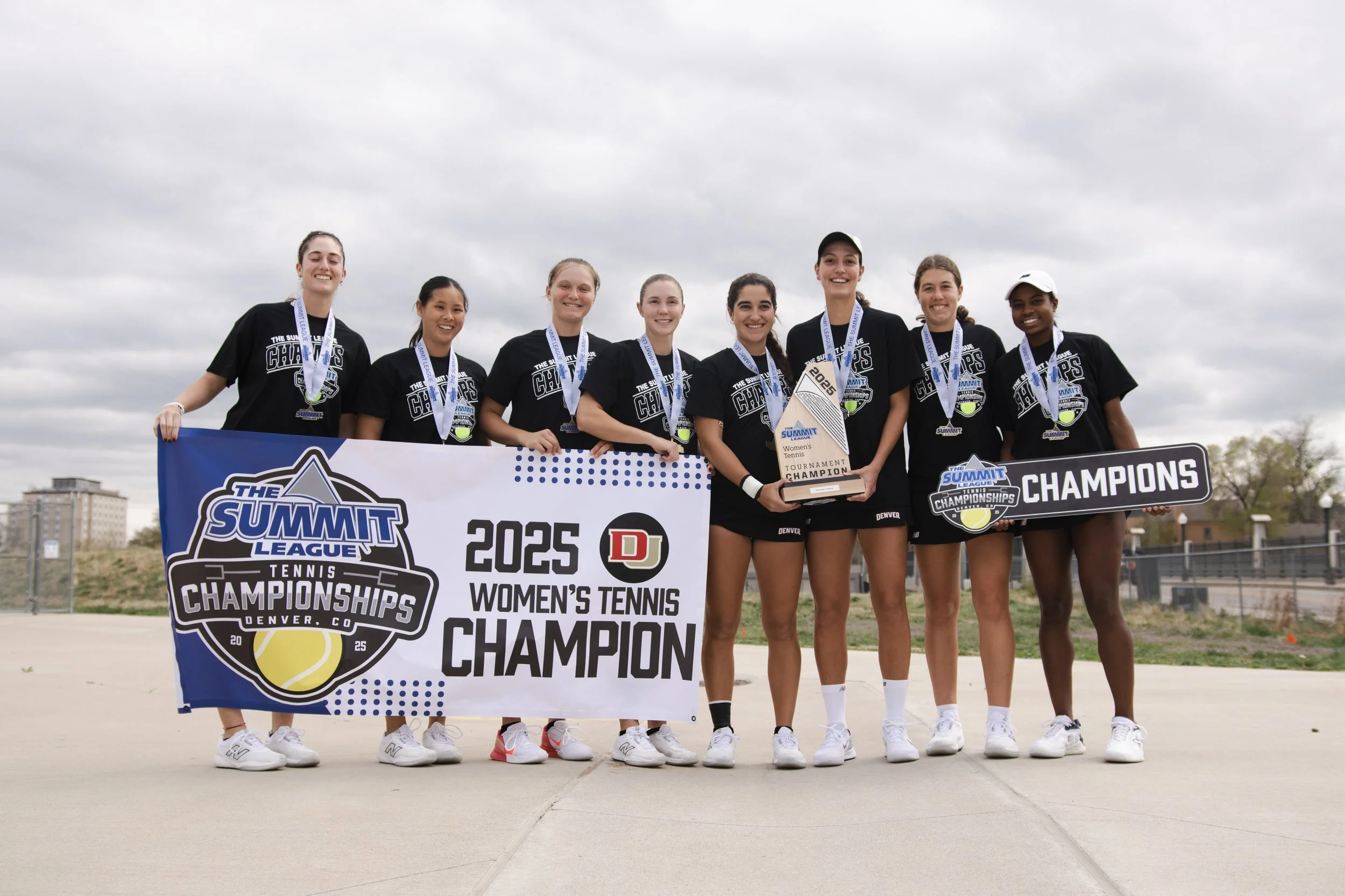 A group of eight young women in black sportswear celebrating on a tennis court, holding a large banner that reads '2025 Women's Tennis Champion' and a trophy, with medals around their necks, after winning a tennis championship in Denver.