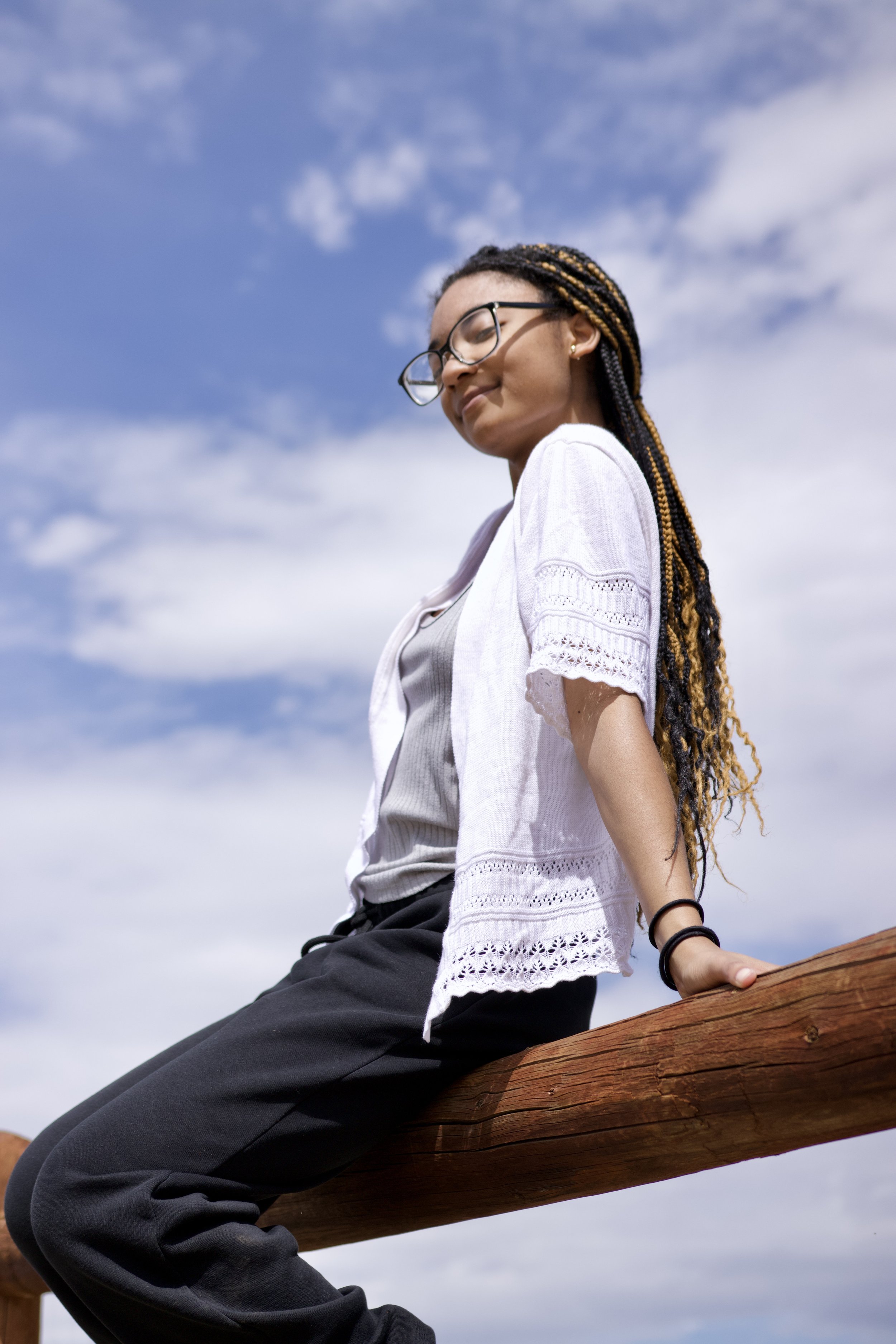 Young woman with glasses sitting on a wooden log outdoors, looking down with a calm expression, under a partly cloudy sky.