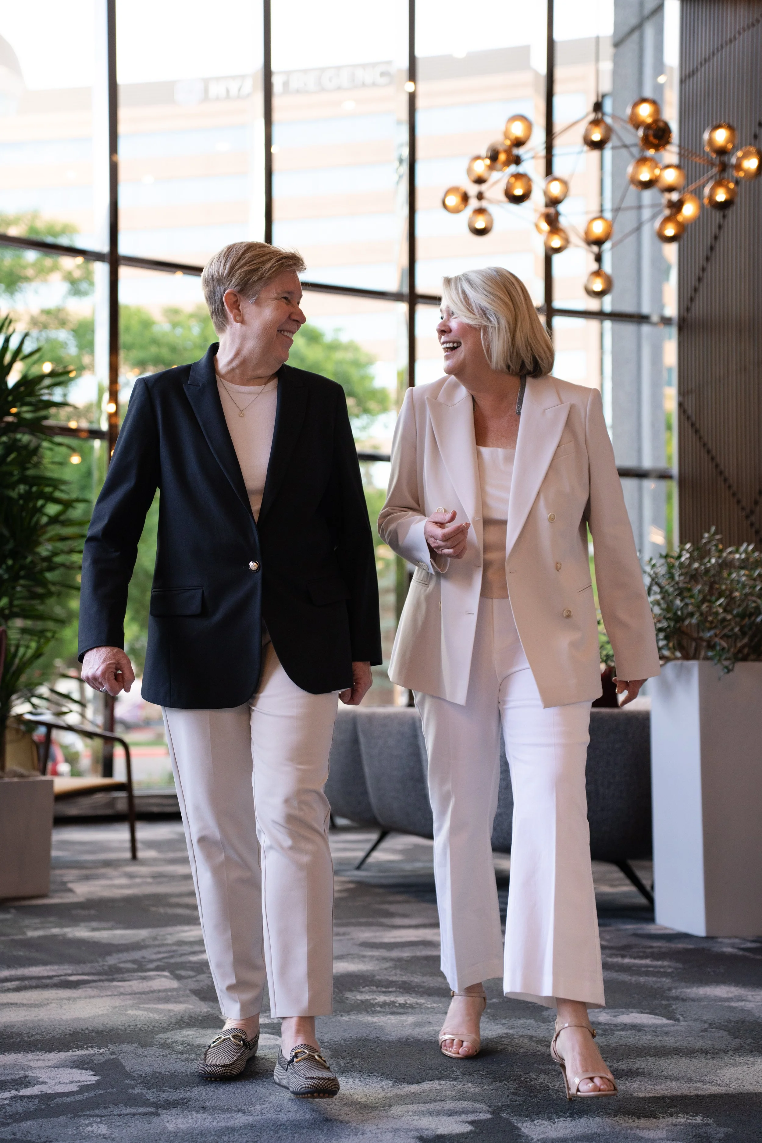 Two women in business attire walking and smiling in a modern office lobby with large windows, plants, and stylish lighting fixtures.