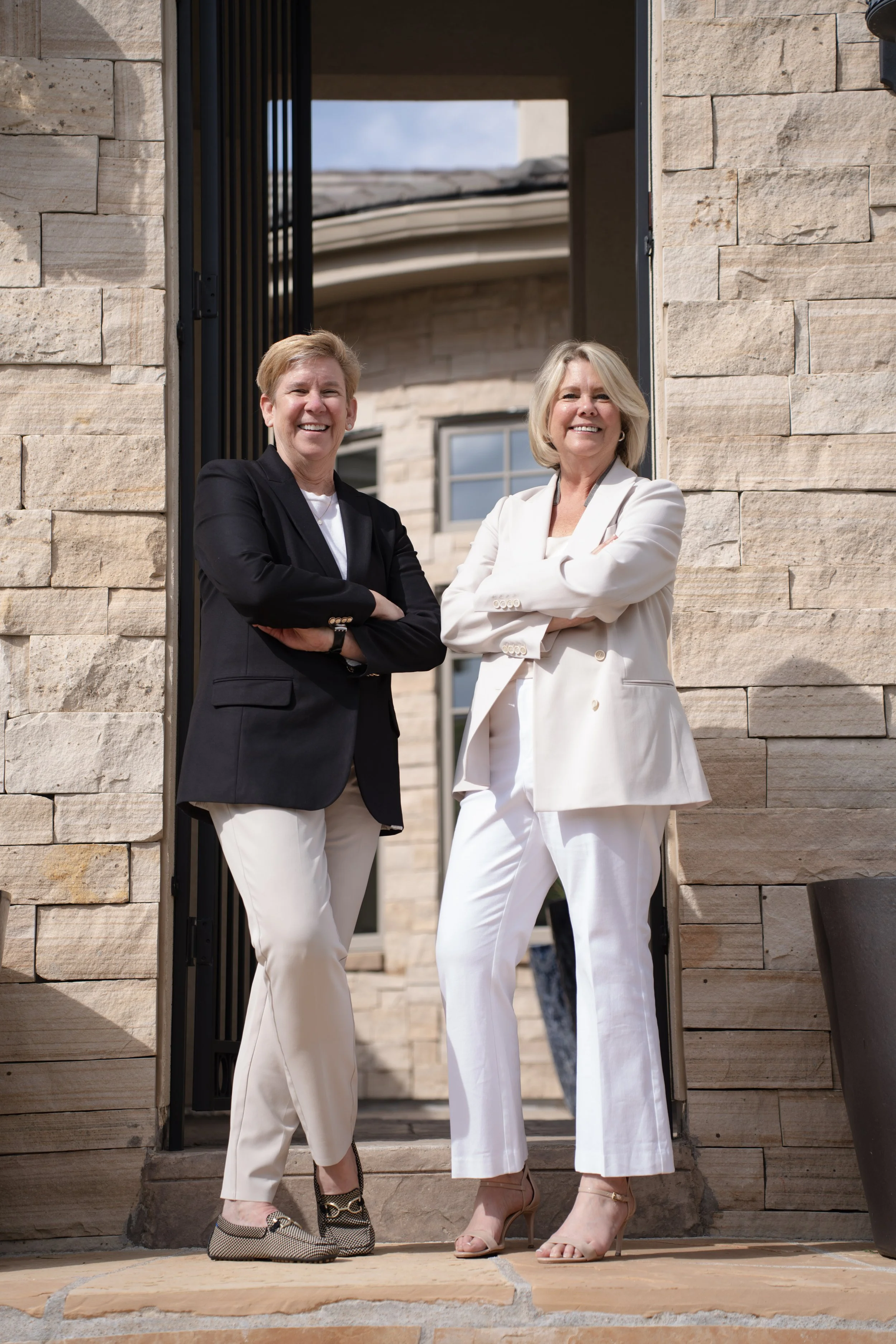 Two professional women standing at the entrance of a building, smiling with arms crossed, dressed in business attire.
