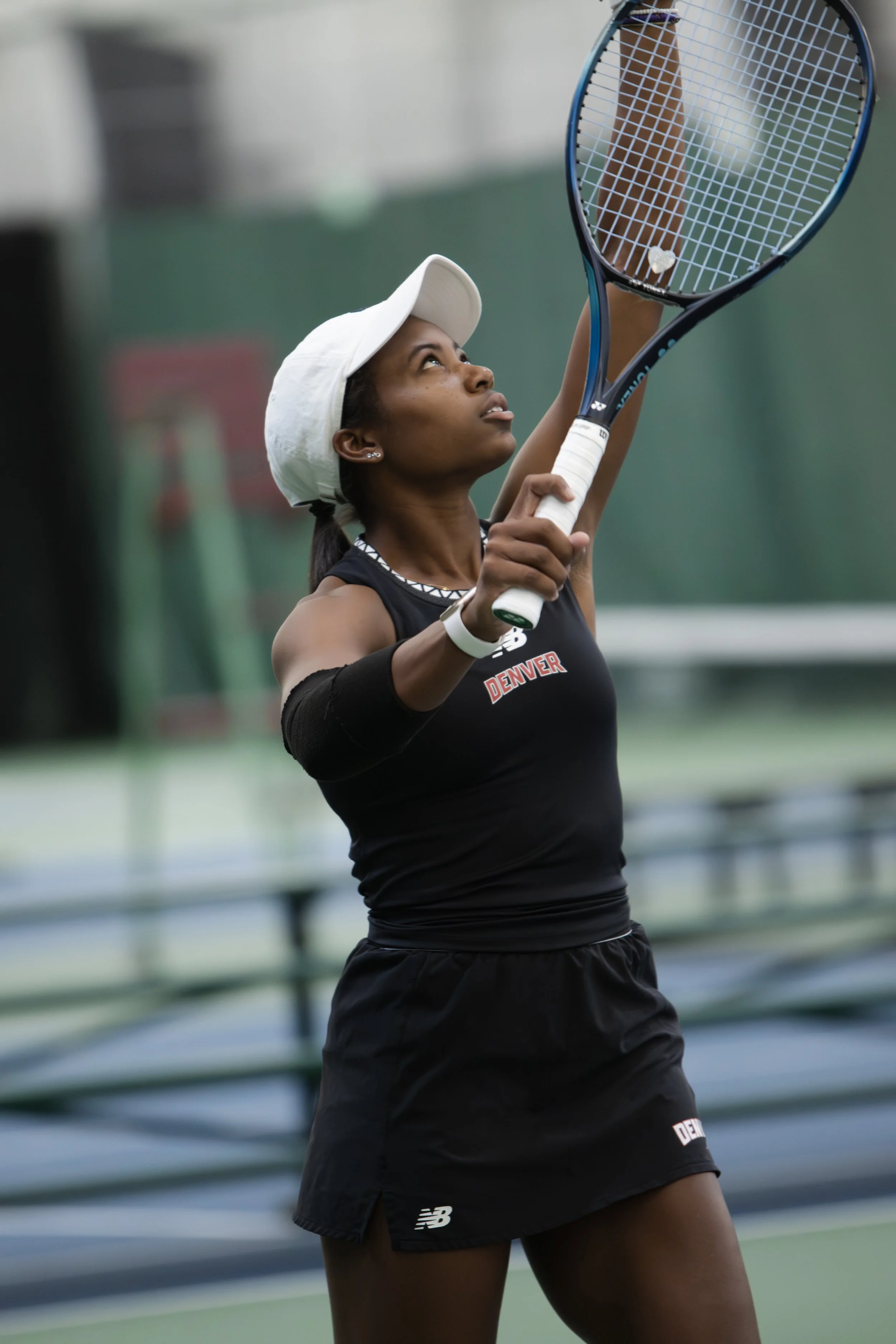 A female tennis player wearing a white cap, black sports attire with 'Denver' on it, and a black armband, about to hit a tennis ball with her racket on an indoor court.