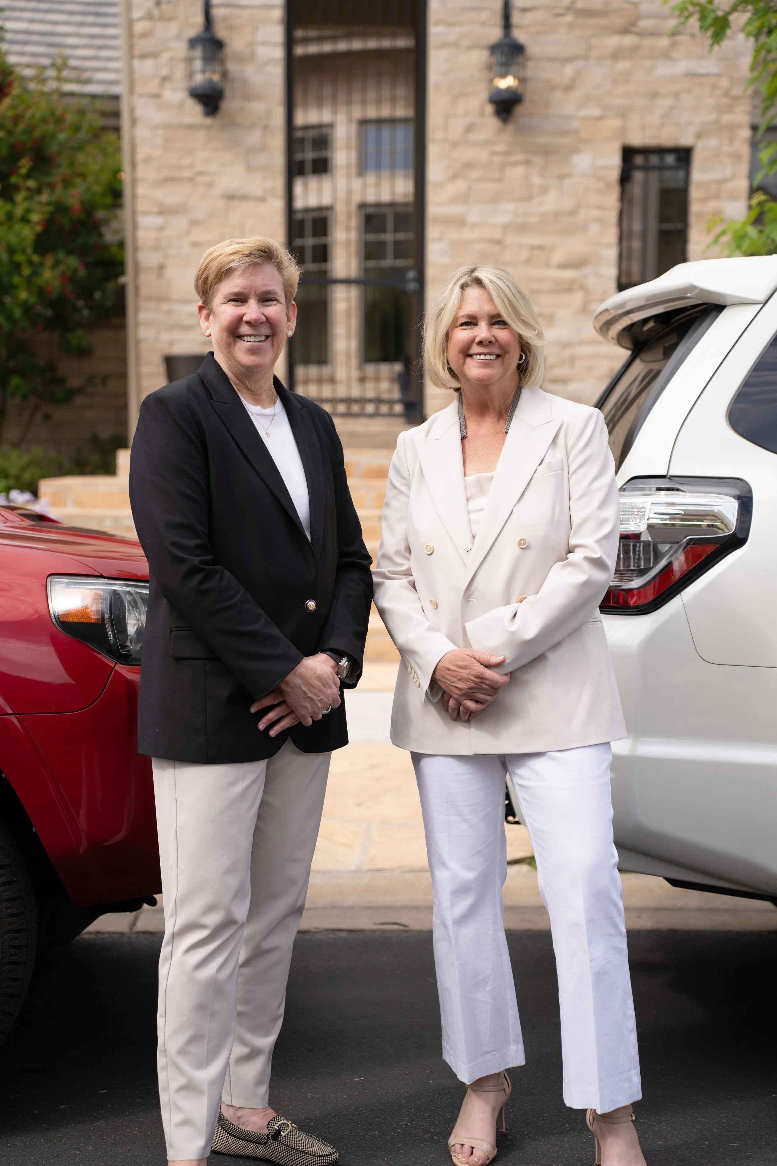 Two women in professional attire smiling and standing outdoors between a red vehicle and a white vehicle in front of a brick building.