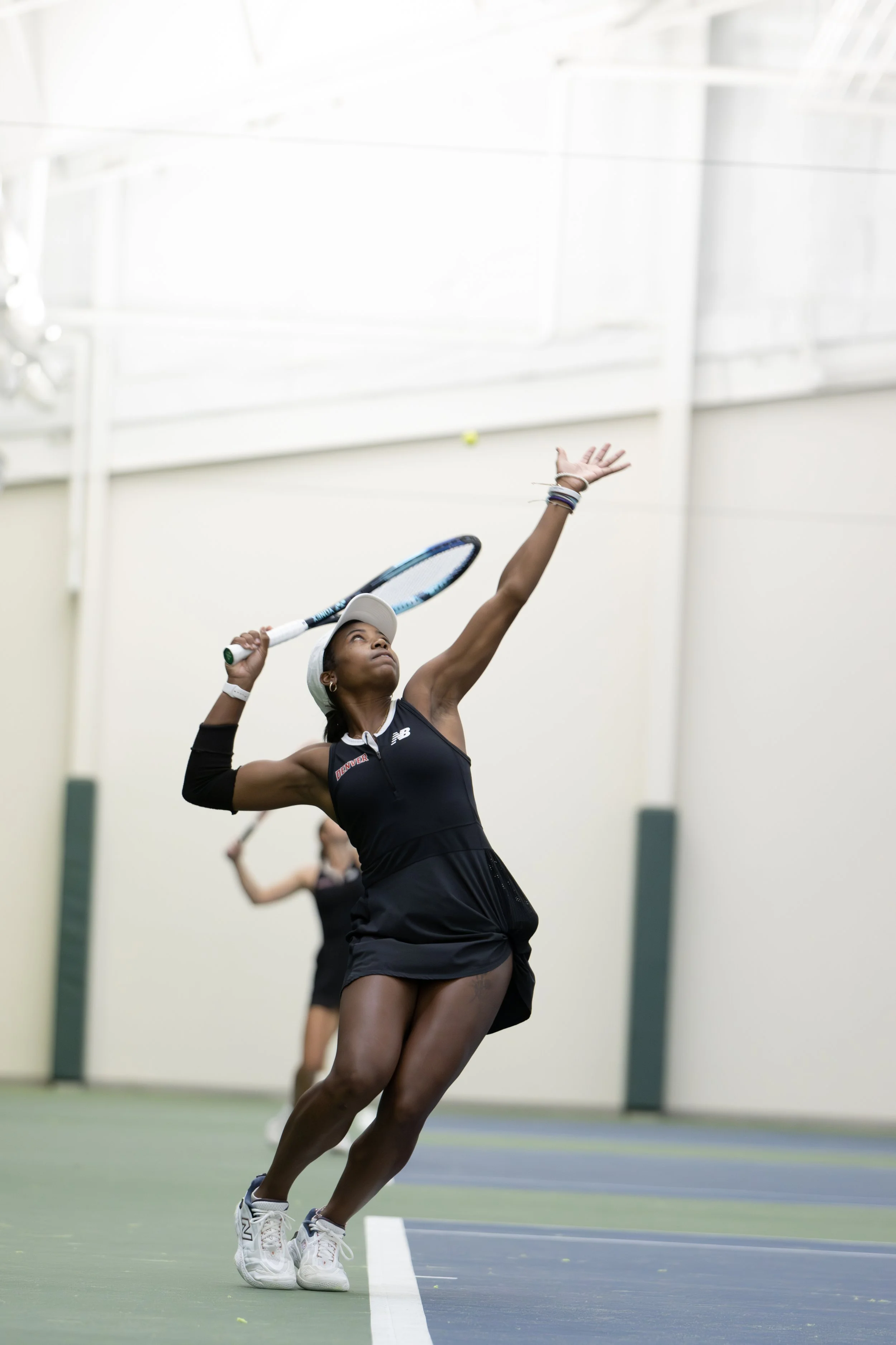 A female tennis player in a black sports dress and white cap serves the ball on an indoor tennis court.