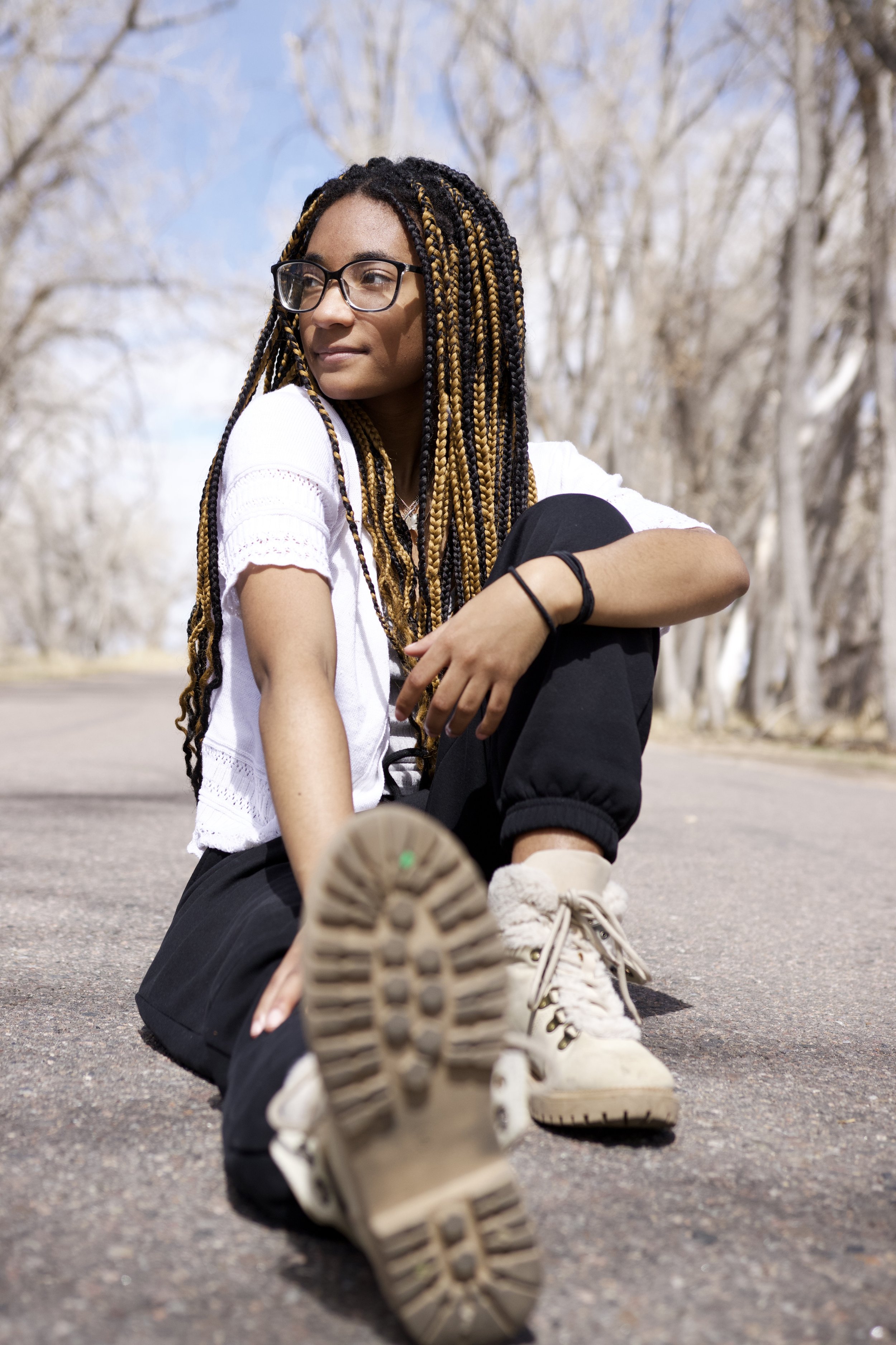 Young woman with glasses and braided hair sitting on the ground outdoors on a paved road, with leafless trees in the background, wearing a white top, black pants, and beige boots.