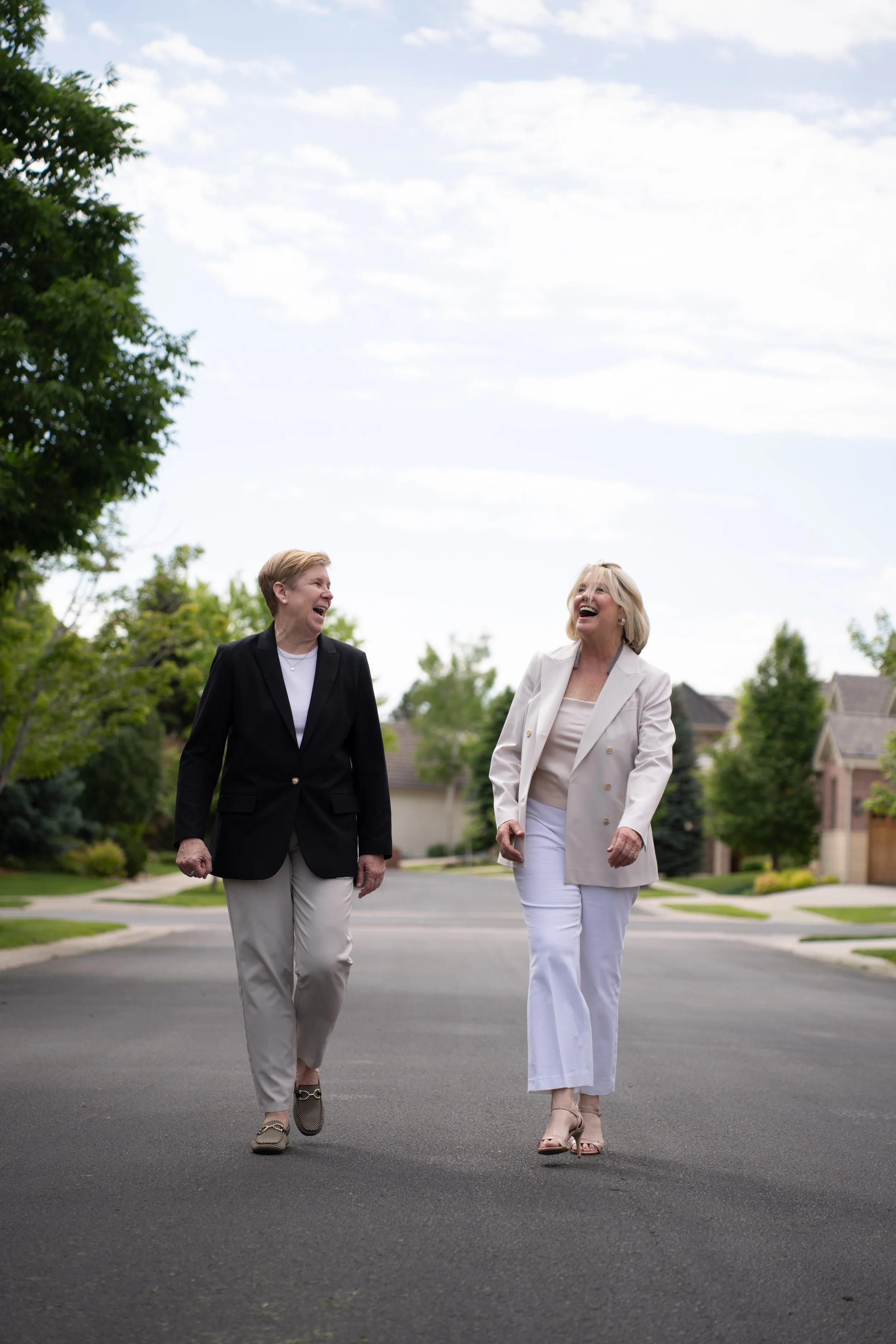 Two women in business attire walking and laughing on a suburban street.