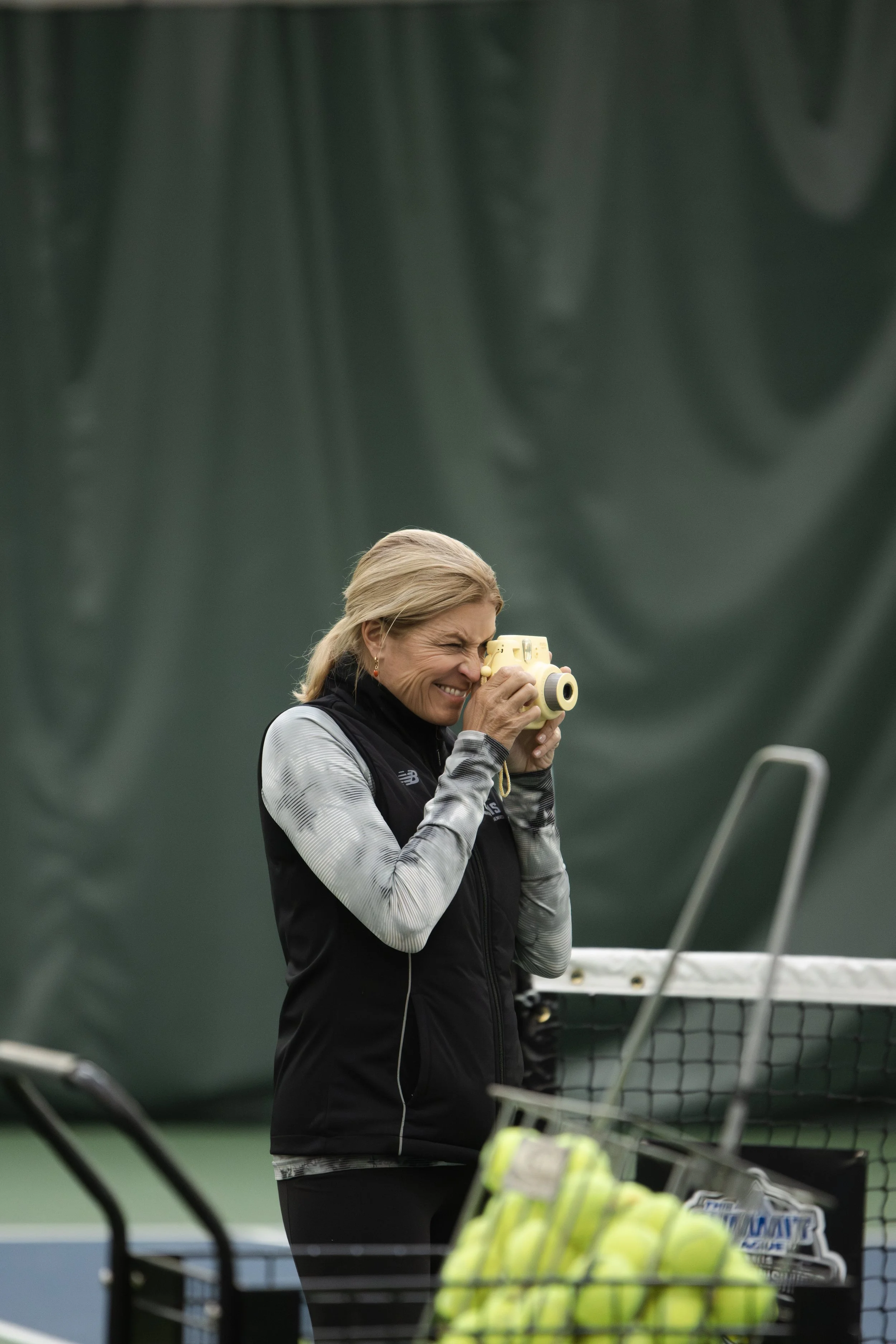 A woman taking a picture with a yellow camera on a tennis court, with tennis balls in a cart in the foreground.