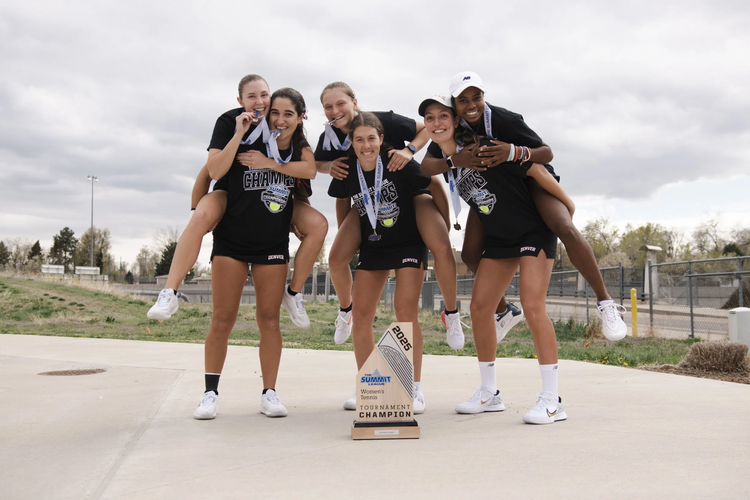 Group of six female tennis players celebrating with medals and a trophy after winning a tournament outdoors on a cloudy day.