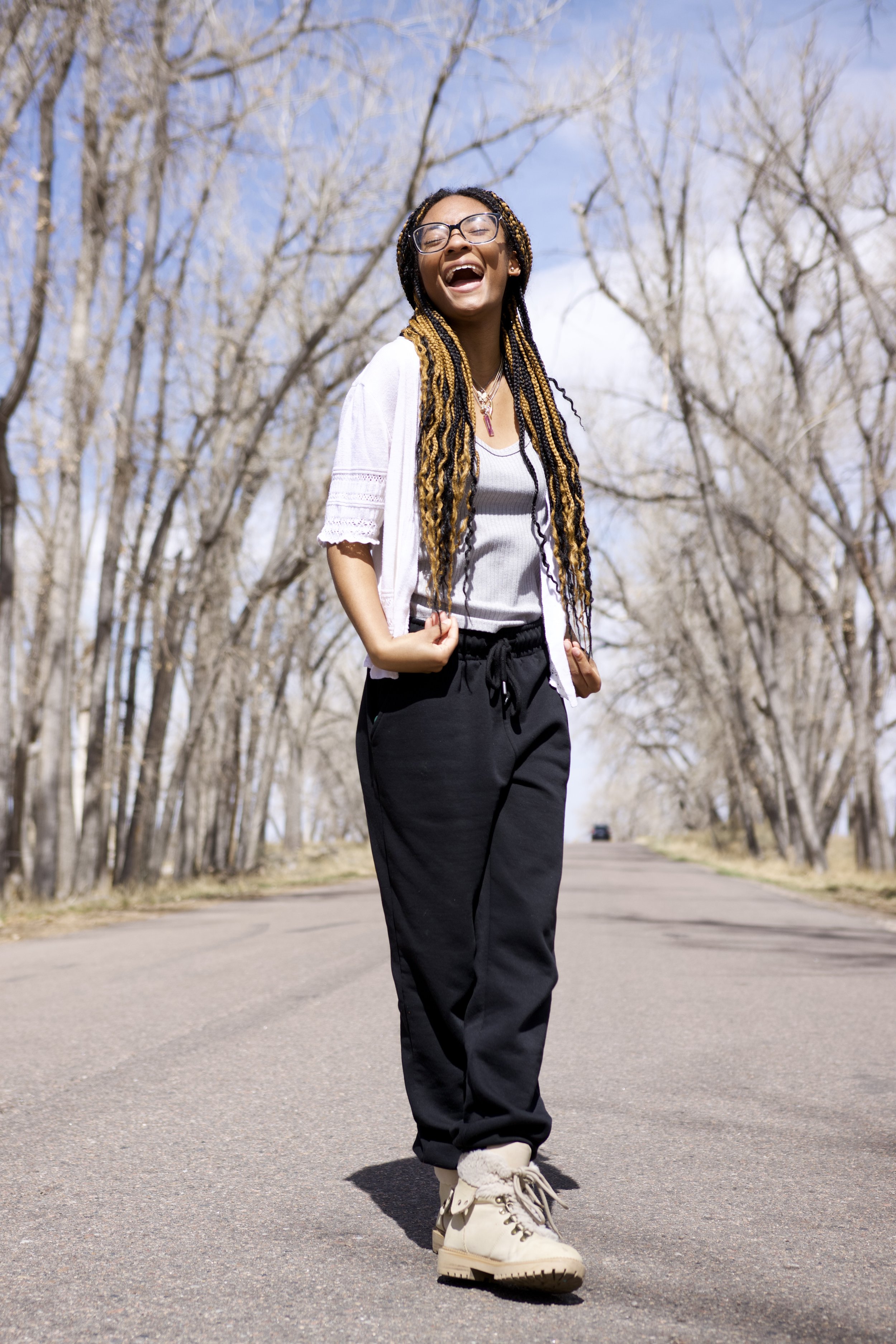 A woman with long braided hair and glasses standing on an empty road, smiling and looking joyful, with leafless trees and a partly cloudy sky in the background.
