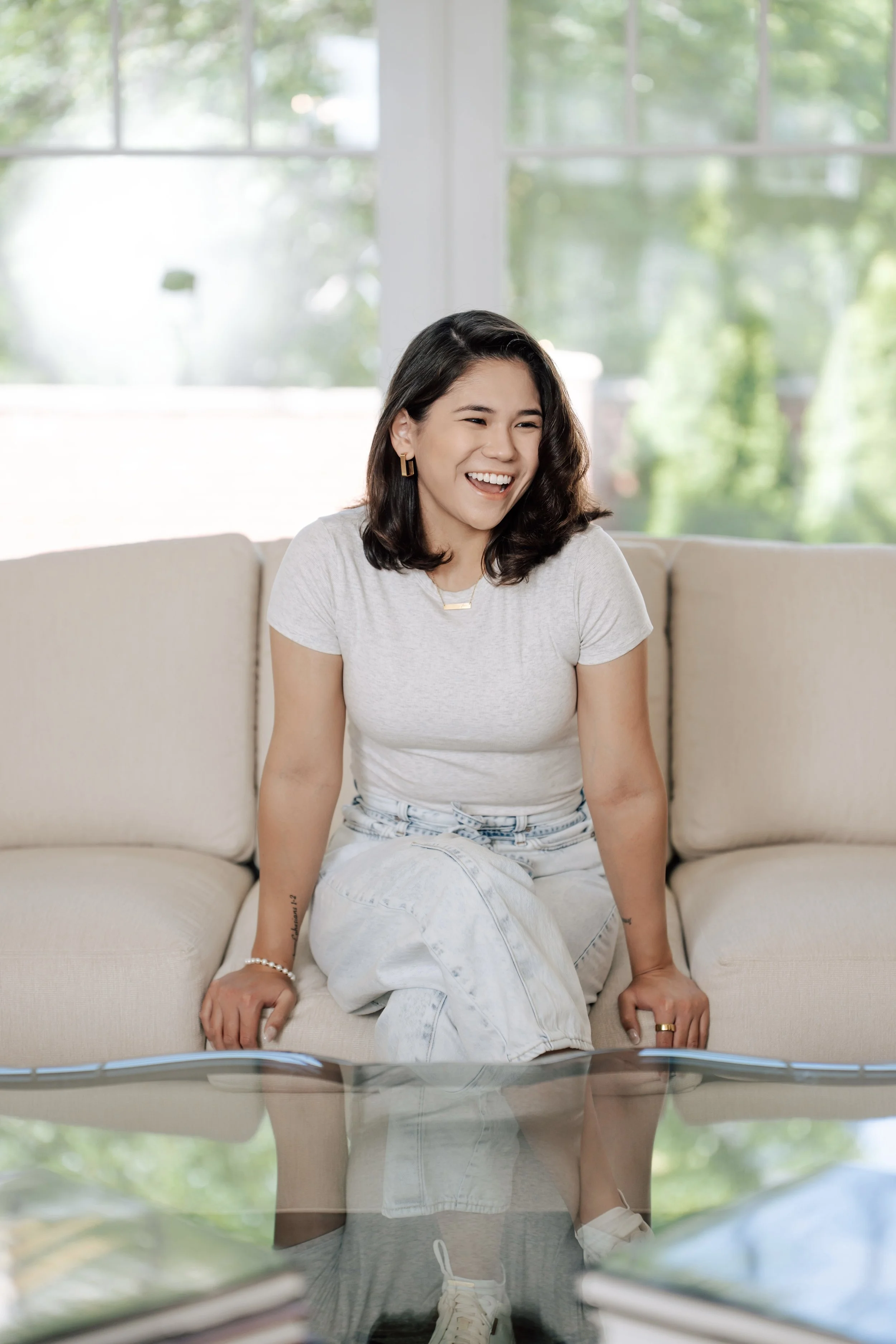 A woman with shoulder-length dark hair, wearing a light grey t-shirt and white jeans, sitting on a beige sofa, smiling and laughing, in front of a glass coffee table with a blurred outdoor background visible through large windows.
