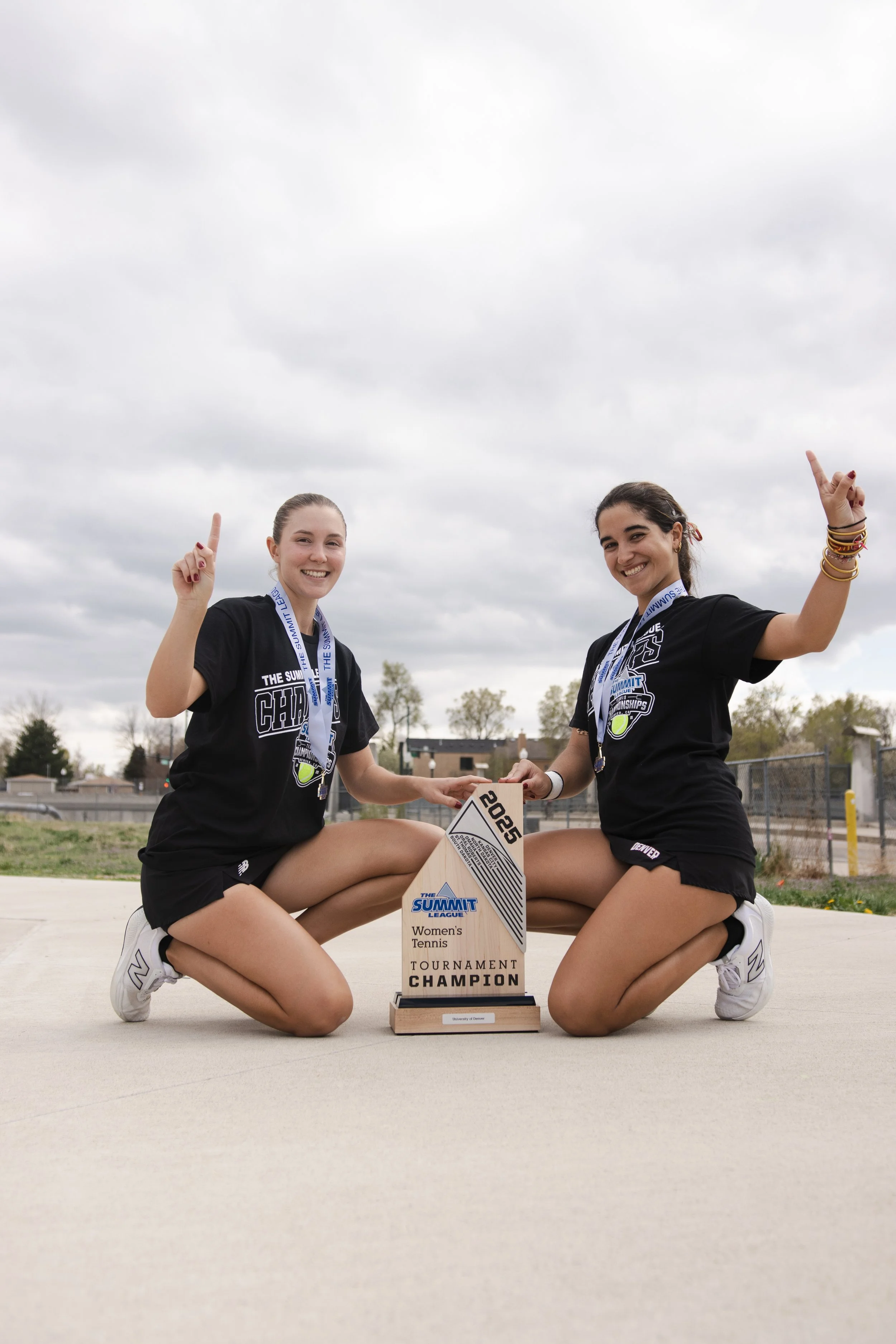 Two female tennis players kneeling on the ground, smiling, holding a tournament championship trophy, wearing medals around their necks, in black sportswear, outdoors with cloudy sky in the background.