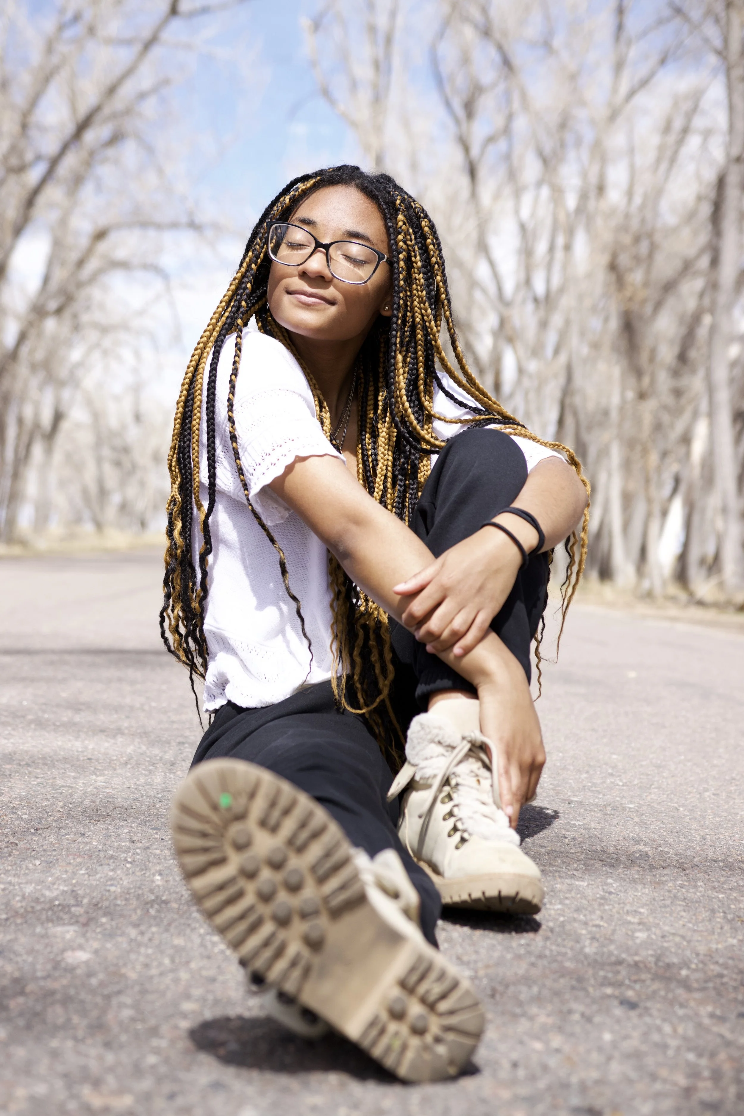 A young woman with long braids, wearing glasses, a white shirt, black pants, and sneakers, sitting on a paved road in a park with leafless trees, enjoying the sunshine with her eyes closed.
