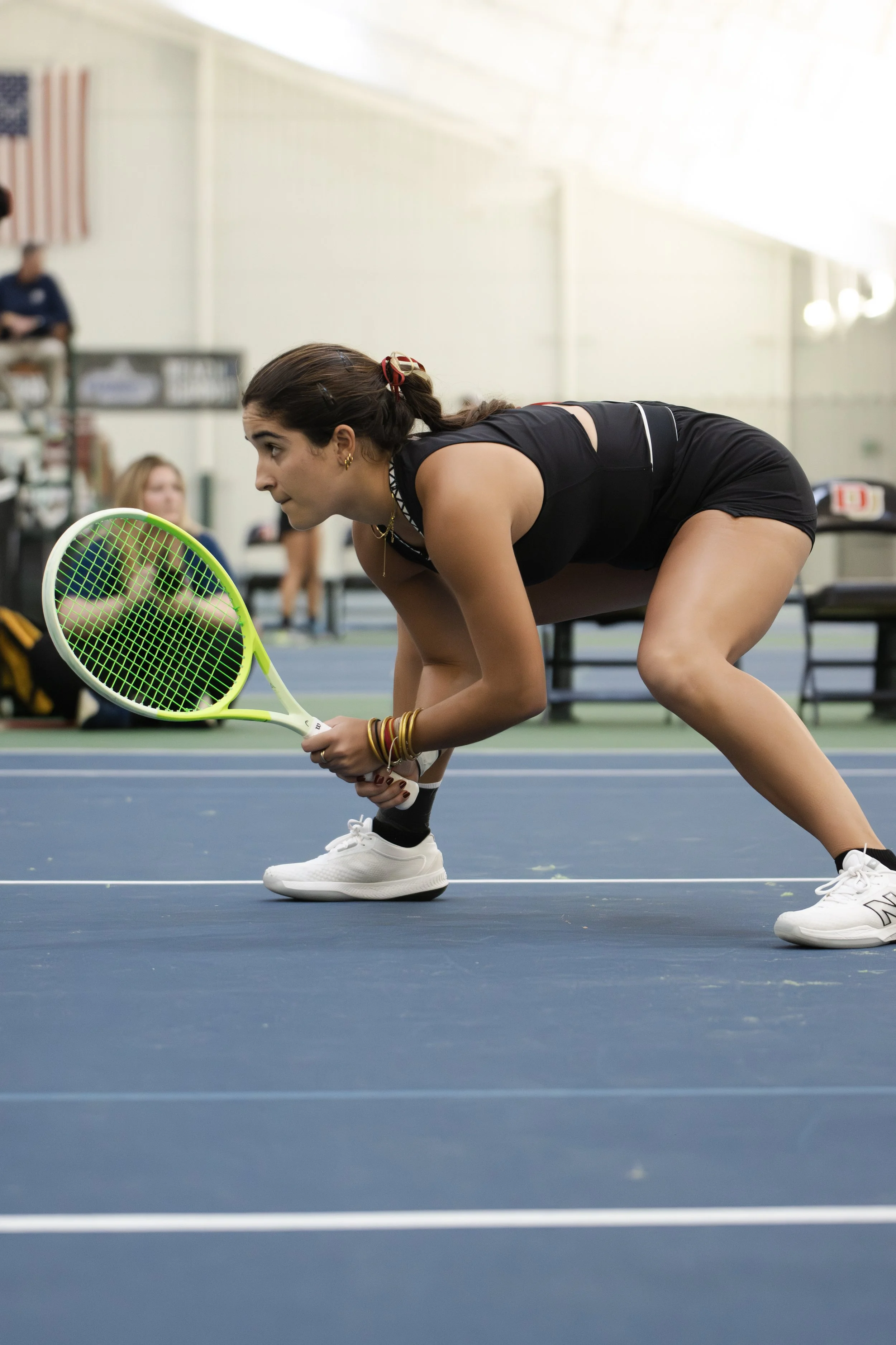 A female tennis player in black athletic attire crouches low to the ground on an indoor tennis court, holding a bright green tennis racket, preparing to make a shot.