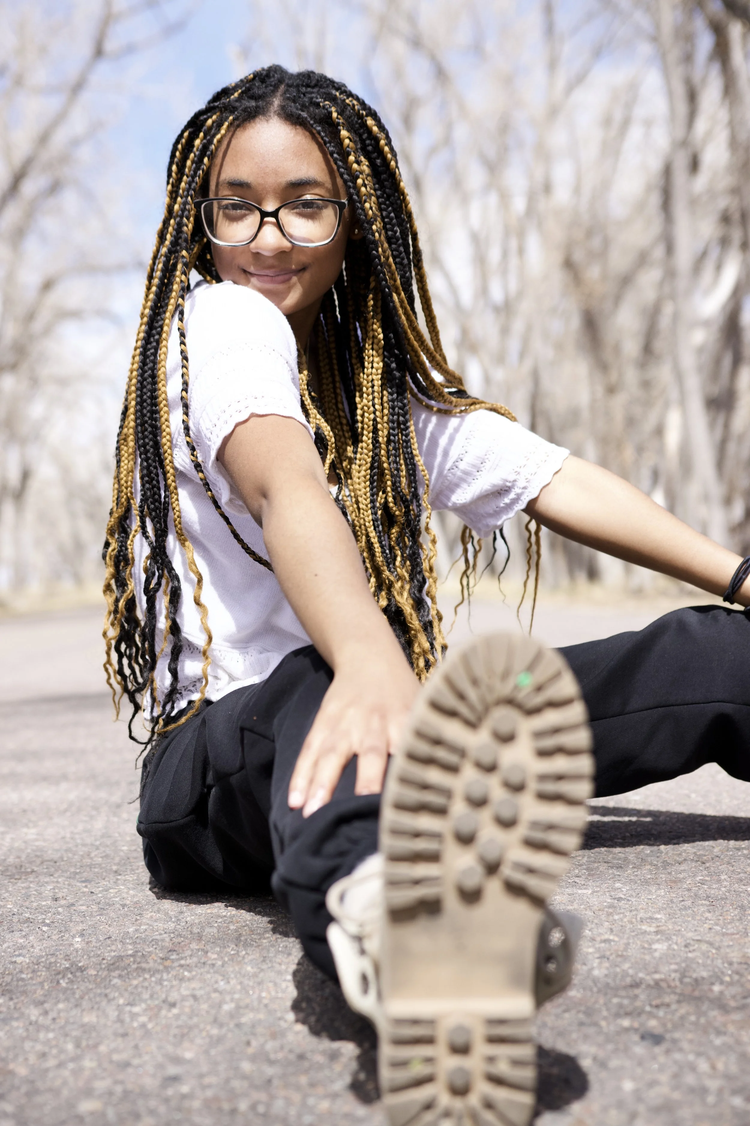 Young woman with glasses and braided hair sitting on pavement outdoors, stretching her leg toward the camera with a background of leafless trees and a clear blue sky.