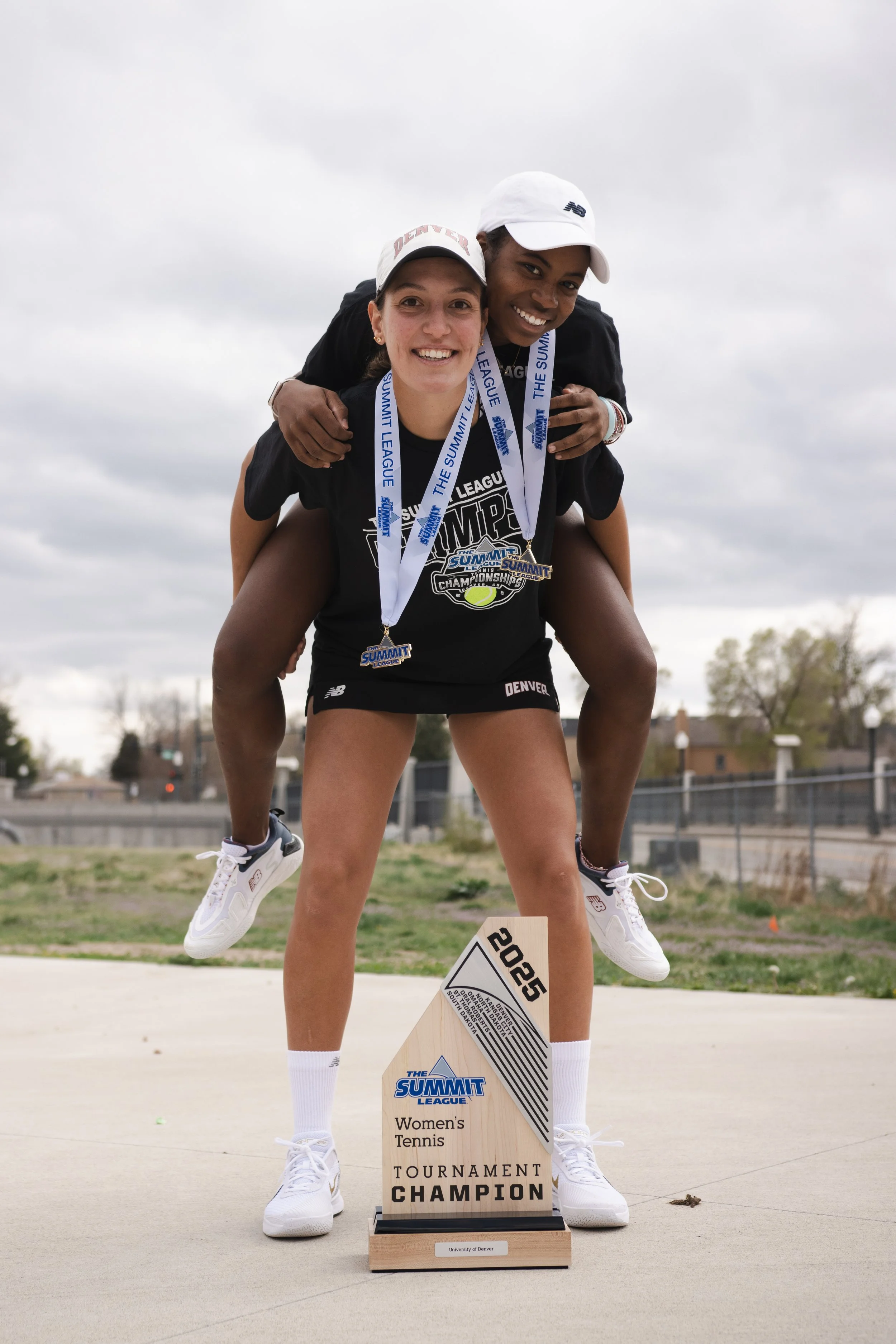 Two female tennis players celebrating as tournament champions, wearing medals around their necks and standing outdoors on a concrete court with a cloudy sky behind them. One player is giving a piggyback ride to the other, and they are smiling.
