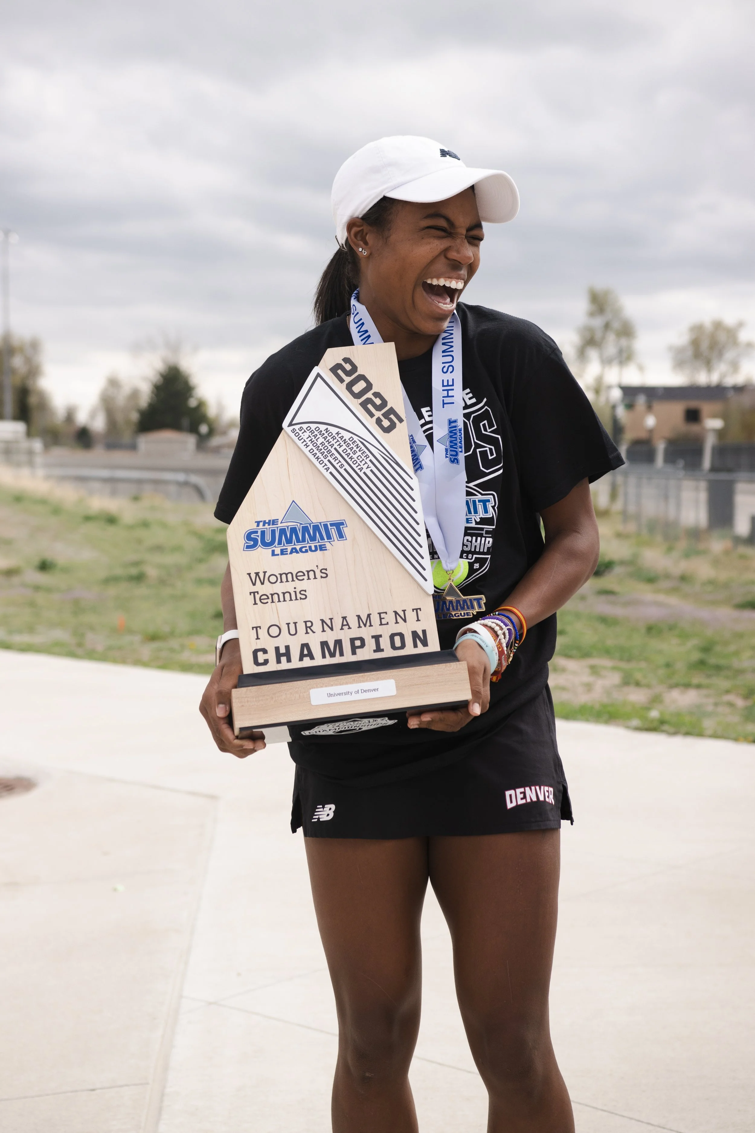 A young woman in athletic attire holding a trophy with the inscription "Women's Tennis Tournament Champion". She is smiling, wearing a cap and medals, celebrating her victory outdoors on a cloudy day.